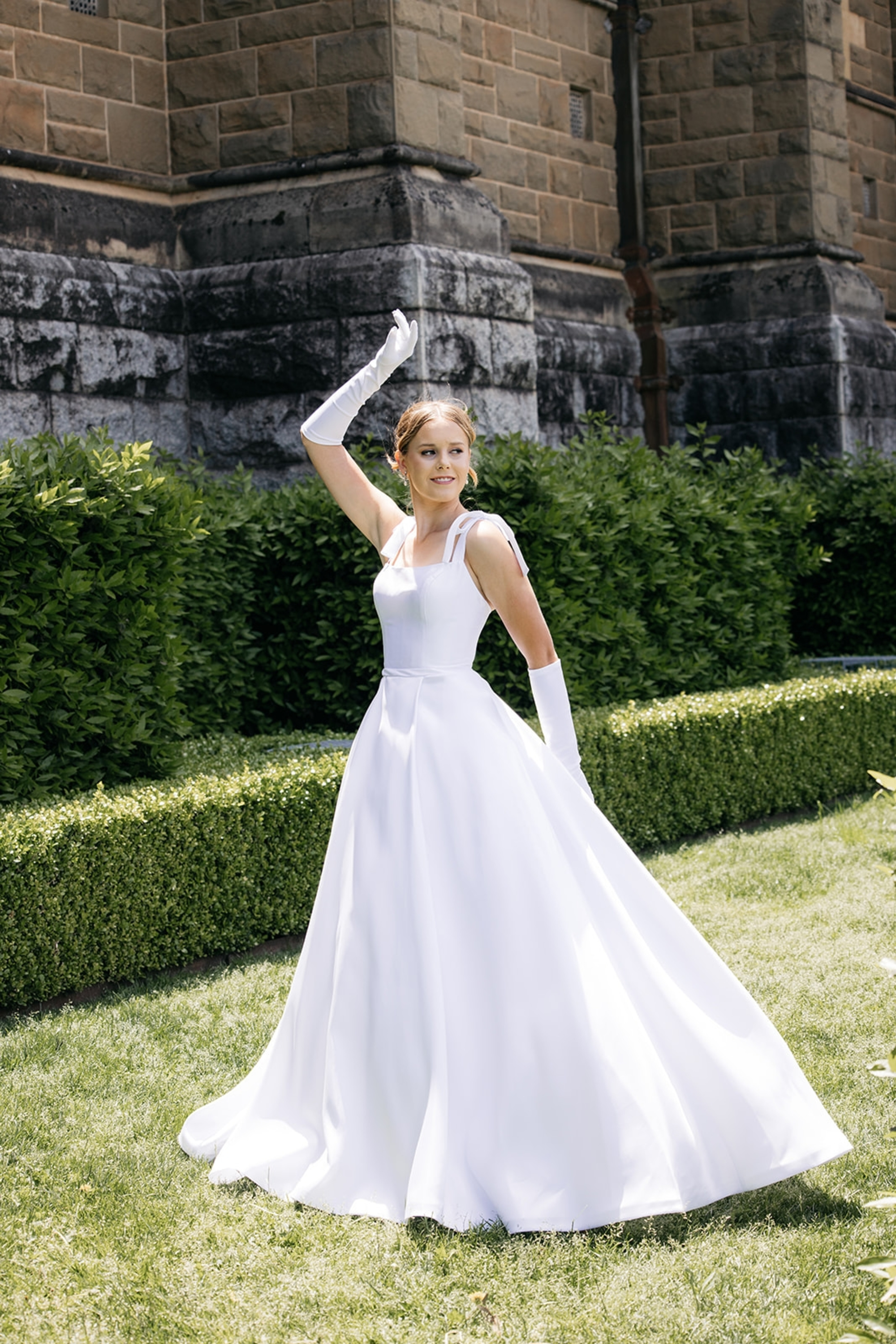 Bride in a classic white gown and gloves posing in a garden outside a stone building.