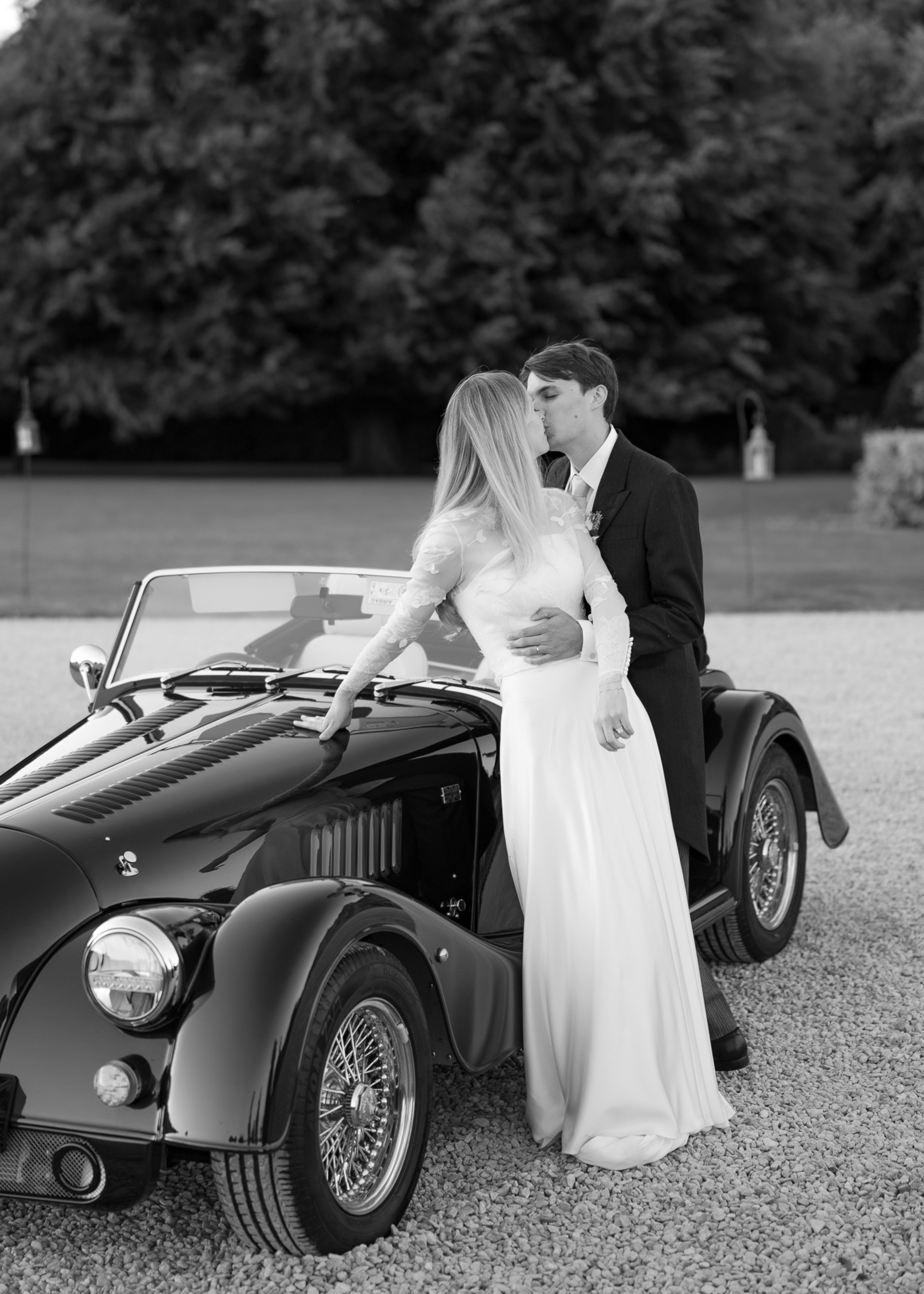 Bride and groom share a kiss beside a classic convertible car in a black and white wedding portrait.