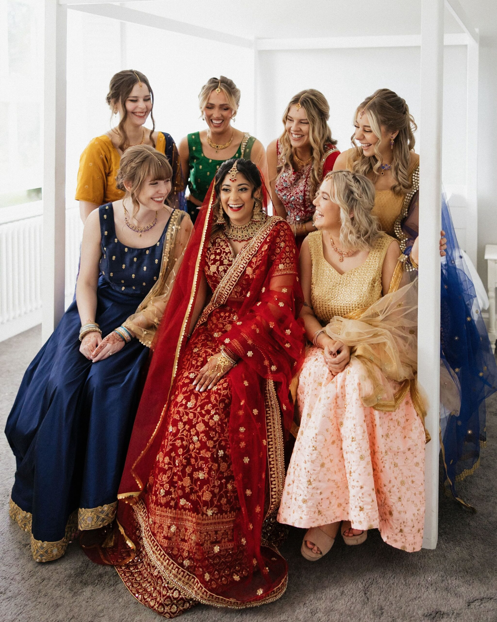 Indian bride in red lehenga smiling with bridesmaids in colorful dresses before the wedding ceremony.