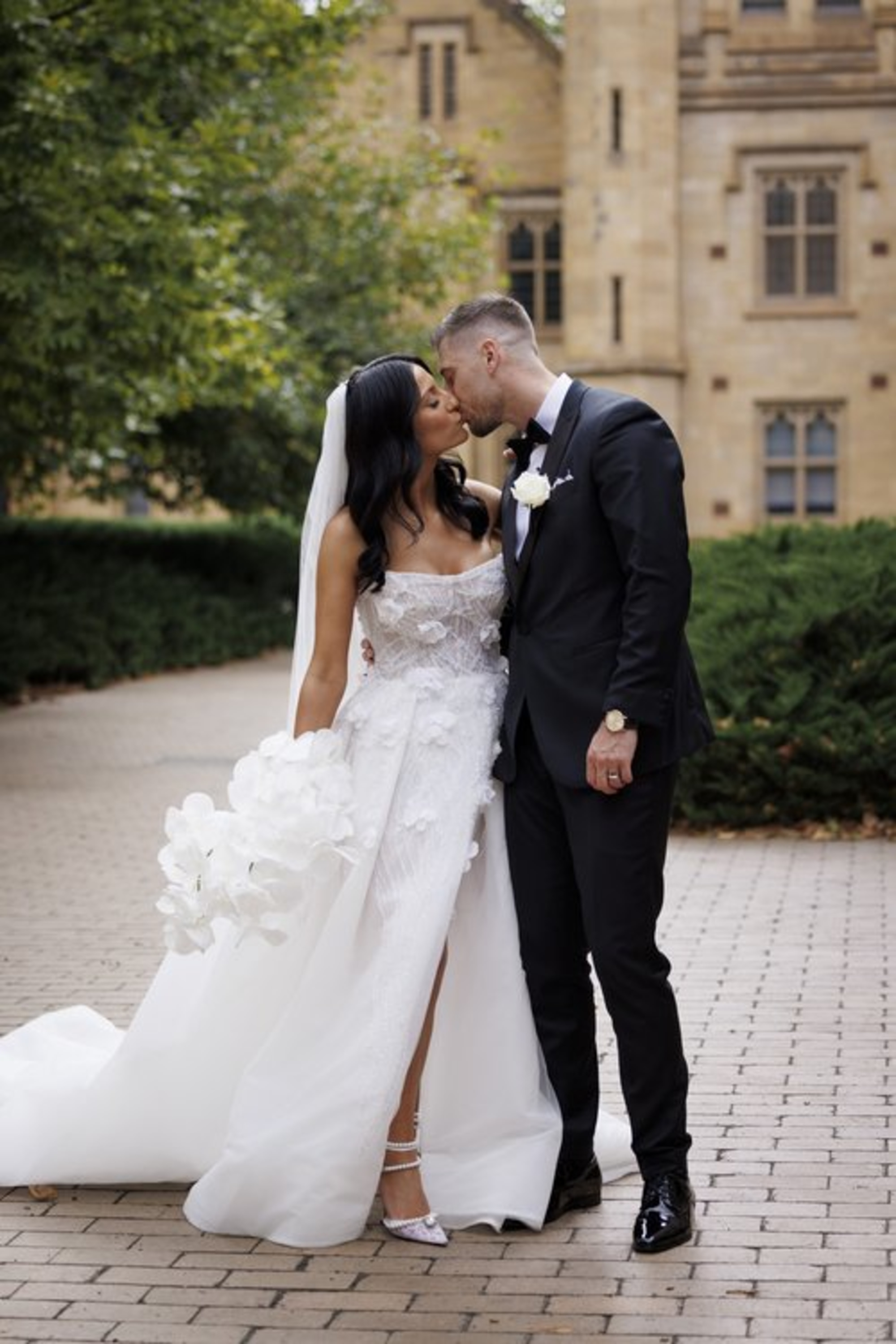 Bride and groom share a kiss outdoors, with the bride in a white gown holding orchids and the groom in a black suit.