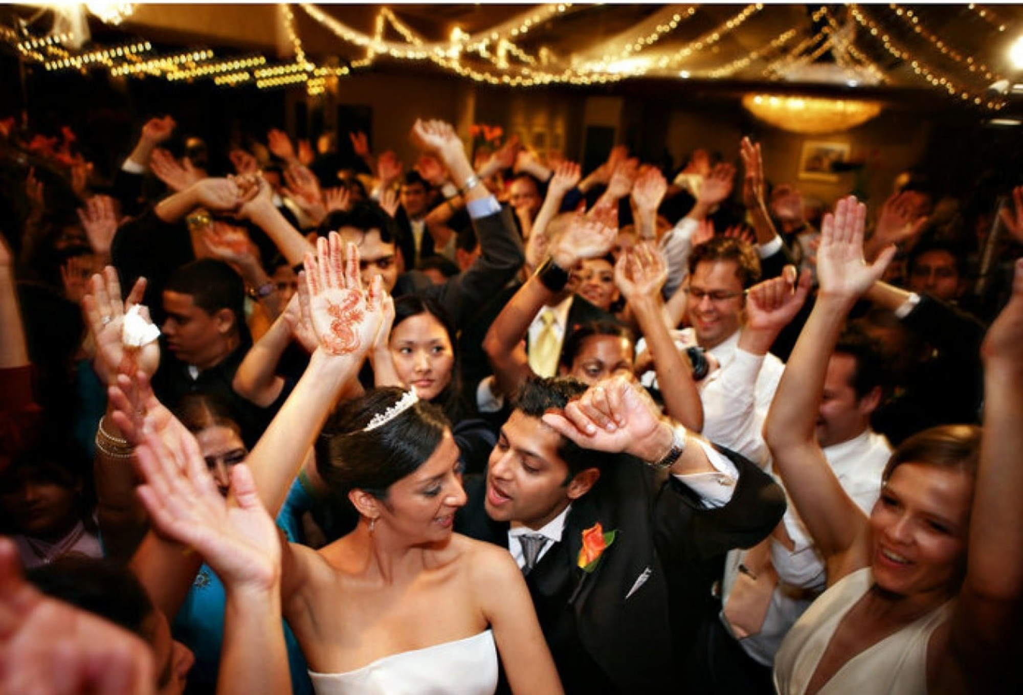 Bride, groom, and guests dancing with raised hands under string lights at a lively wedding reception.
