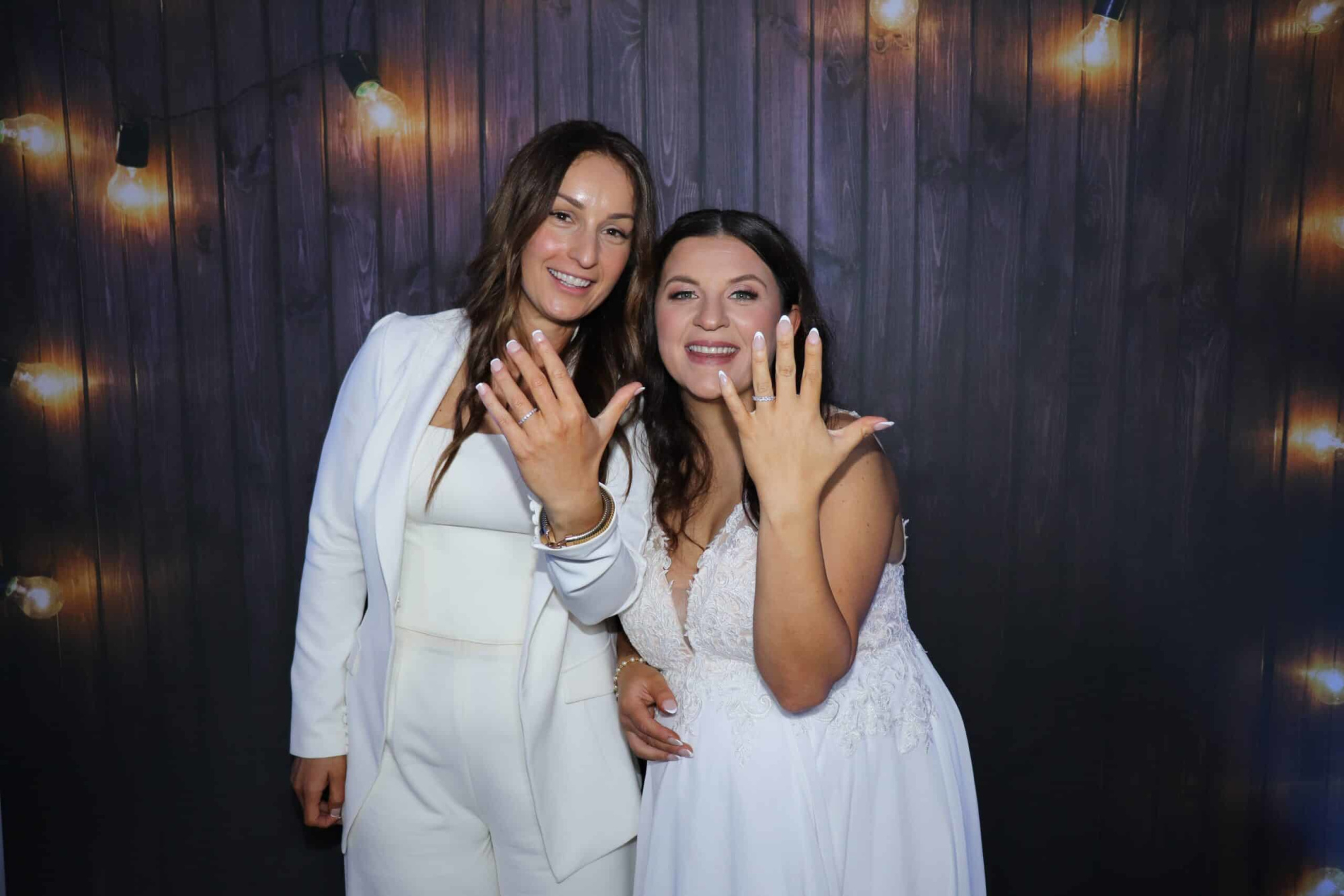 Two newlywed brides pose in front of string lights, smiling and showing off their wedding rings.