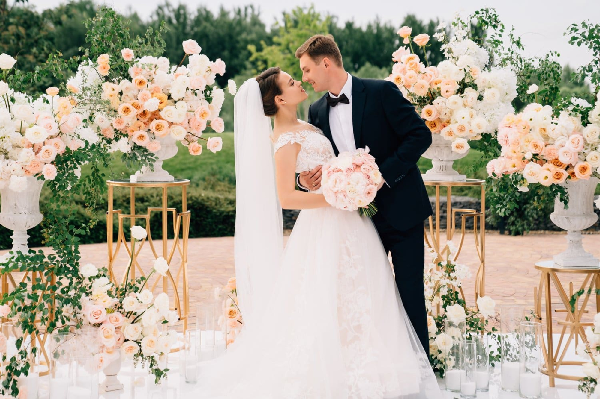 Bride and groom embrace outdoors surrounded by lush peach and white floral arrangements on gold stands.