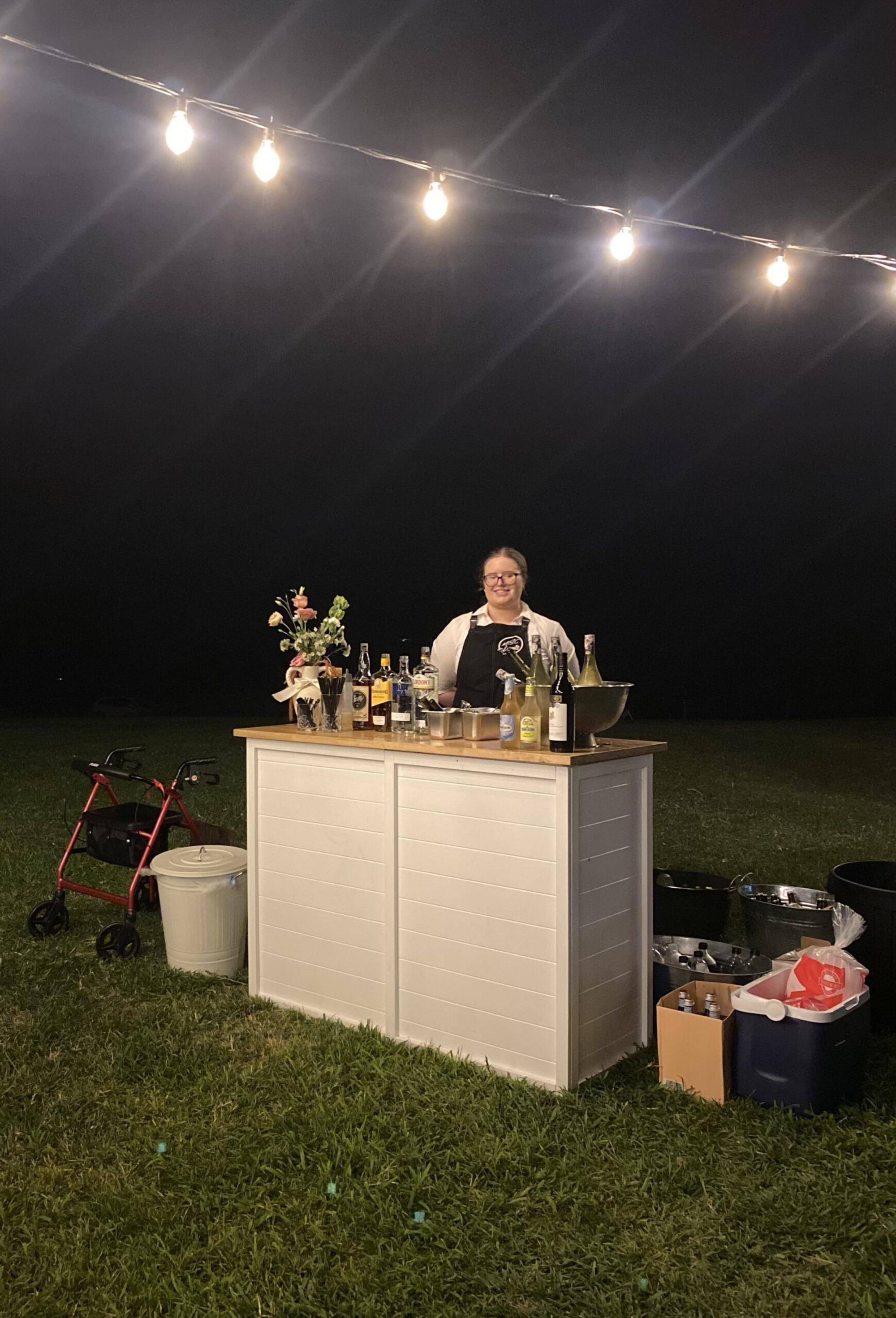 Smiling bartender stands behind a stocked outdoor bar under string lights at a nighttime wedding reception.