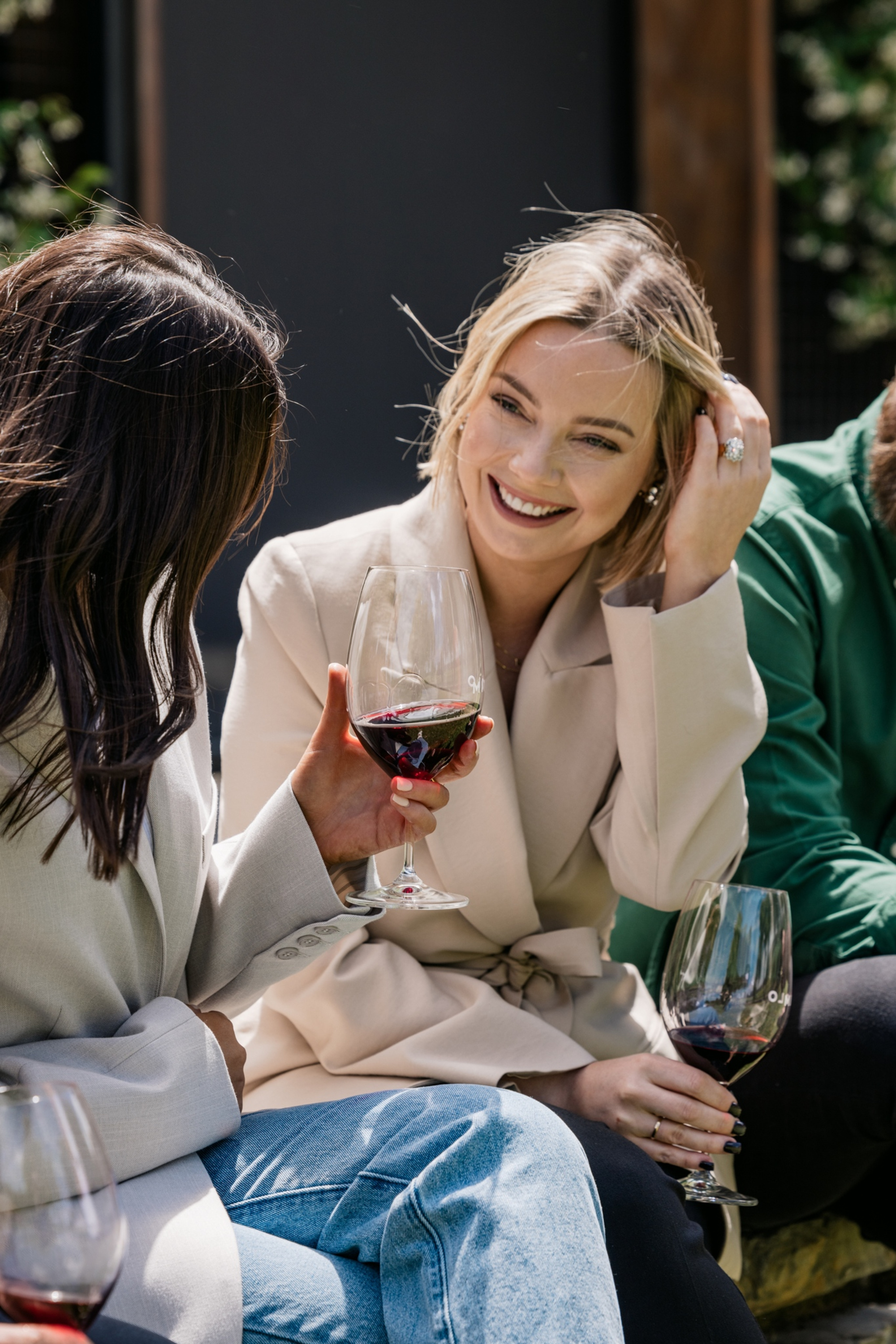 Smiling guests in blazers enjoy glasses of red wine at an outdoor wedding celebration.
