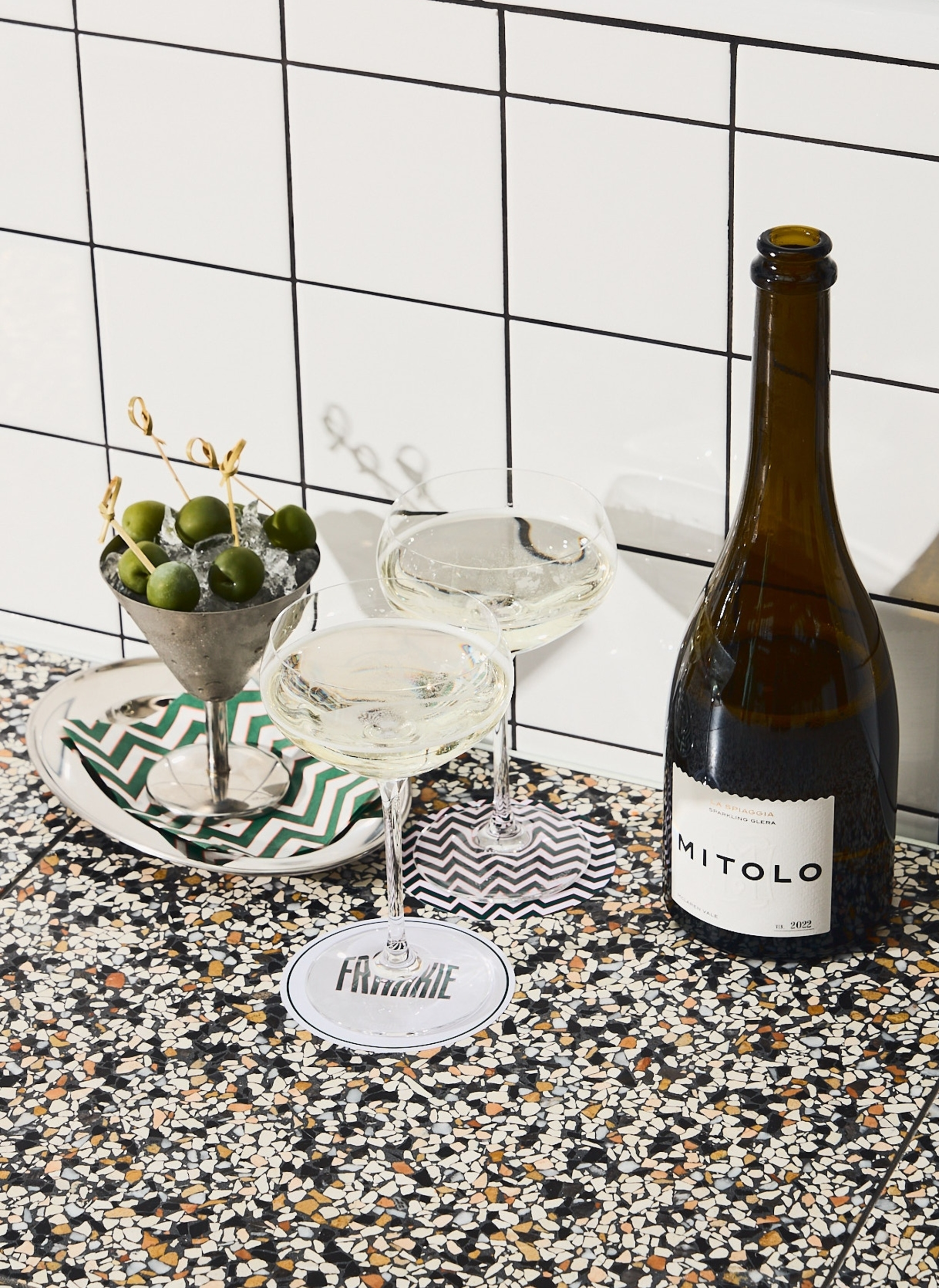 Champagne bottle with two coupe glasses and skewered olives on a terrazzo bar against white tile.