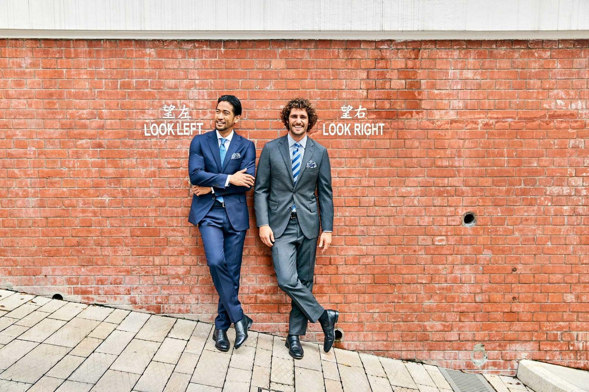 Two grooms in suits lean against a red brick wall with look left and look right signs in an urban setting.