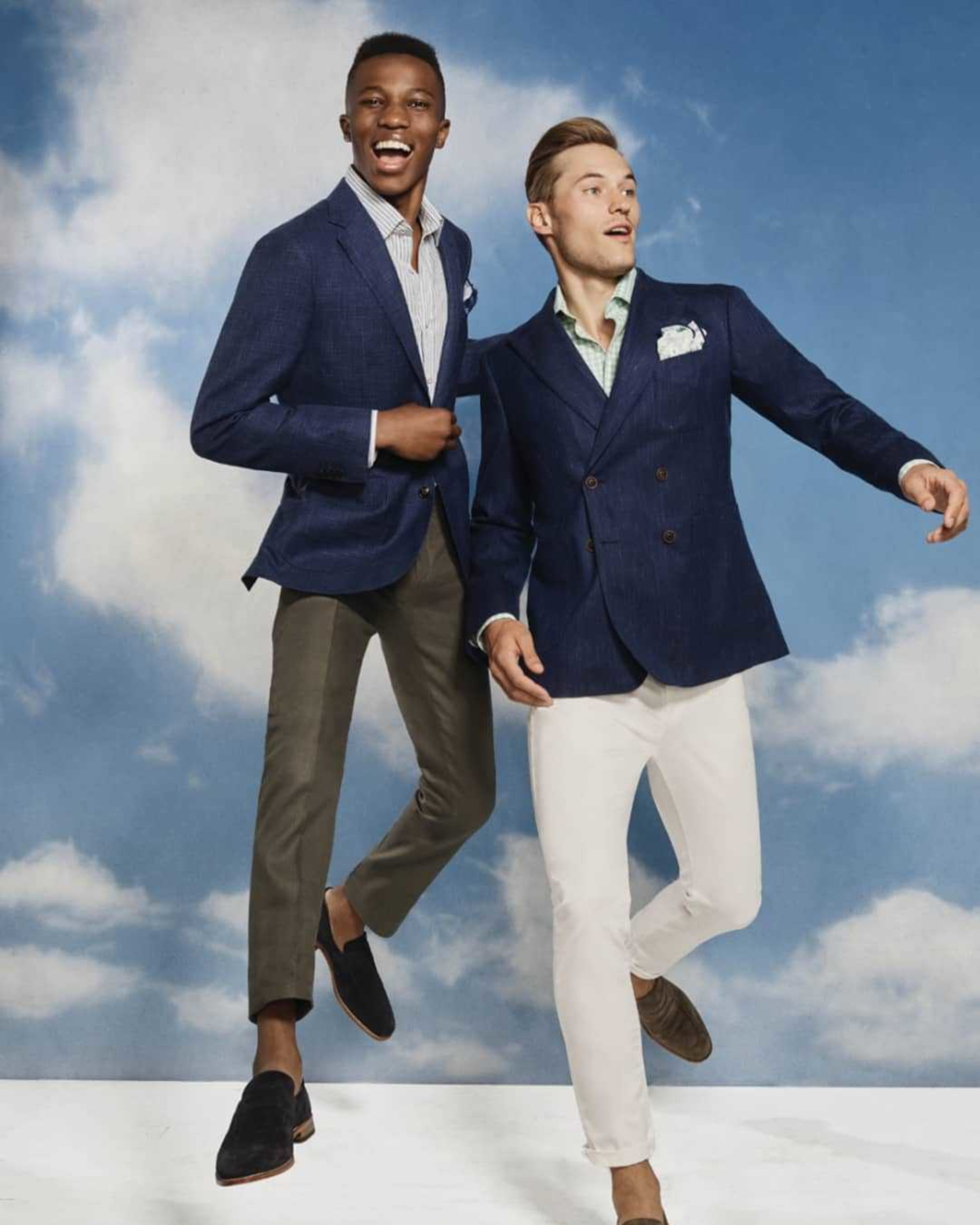 Two men in stylish navy blazers and slim trousers pose against a blue sky backdrop.