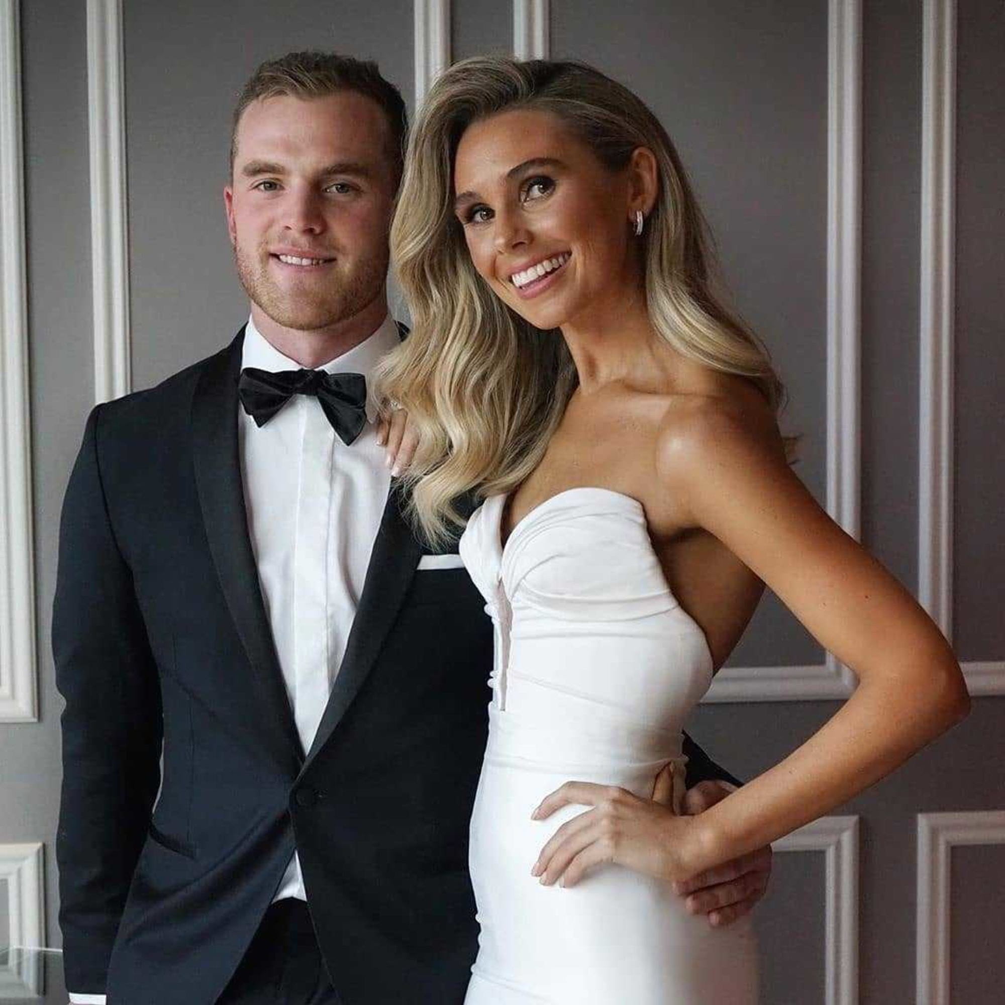 Smiling bride in a strapless white gown posing with groom in a black tuxedo against a paneled wall.