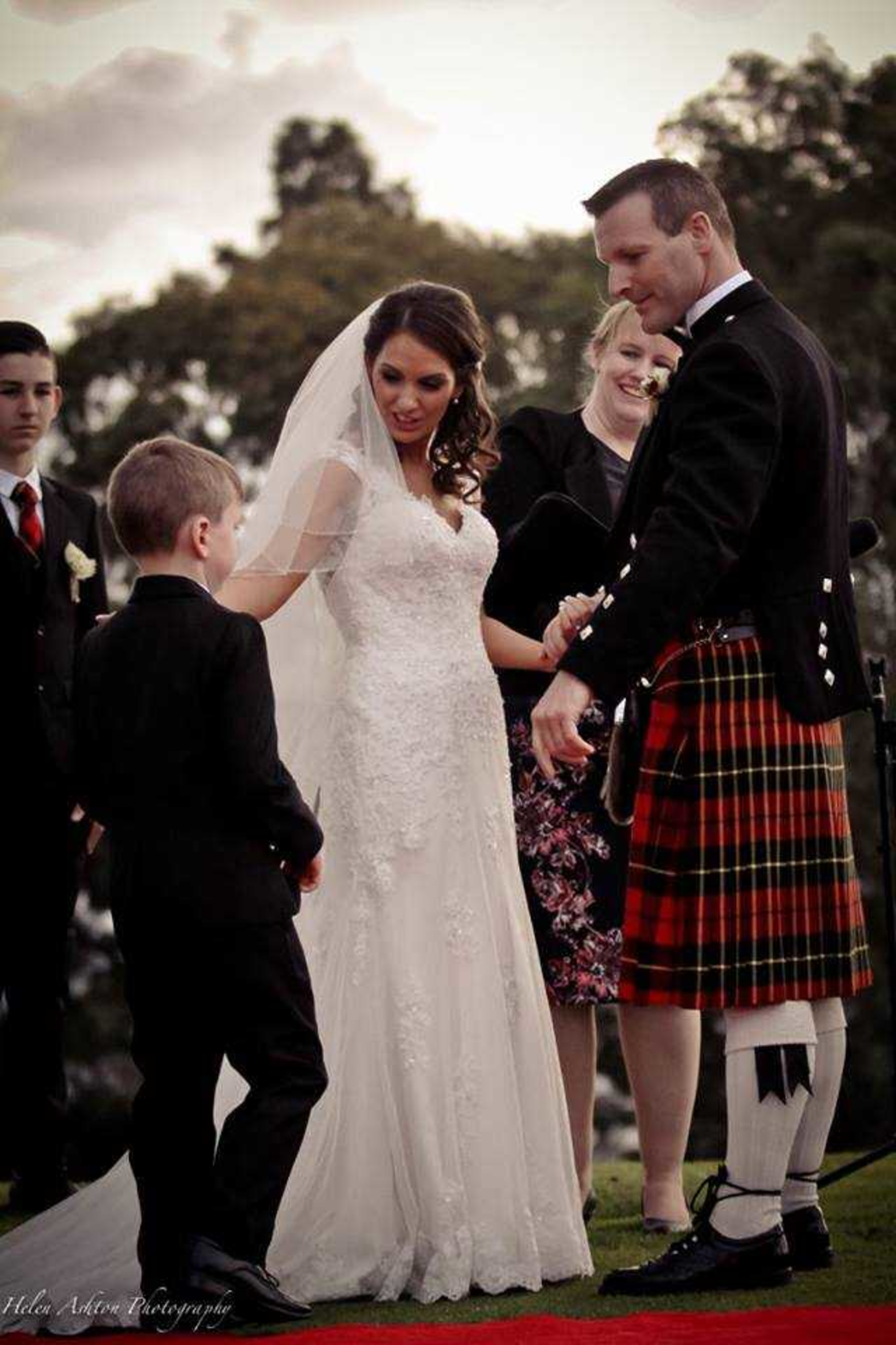 Bride and groom in a tartan kilt share a sweet outdoor ceremony moment with a young boy on the lawn.