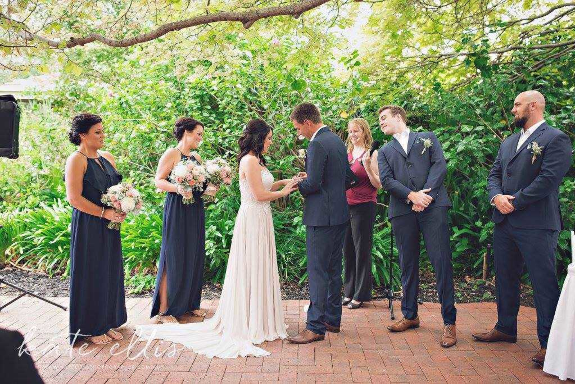 Bride and groom exchange rings during an outdoor garden ceremony with their wedding party beside them.