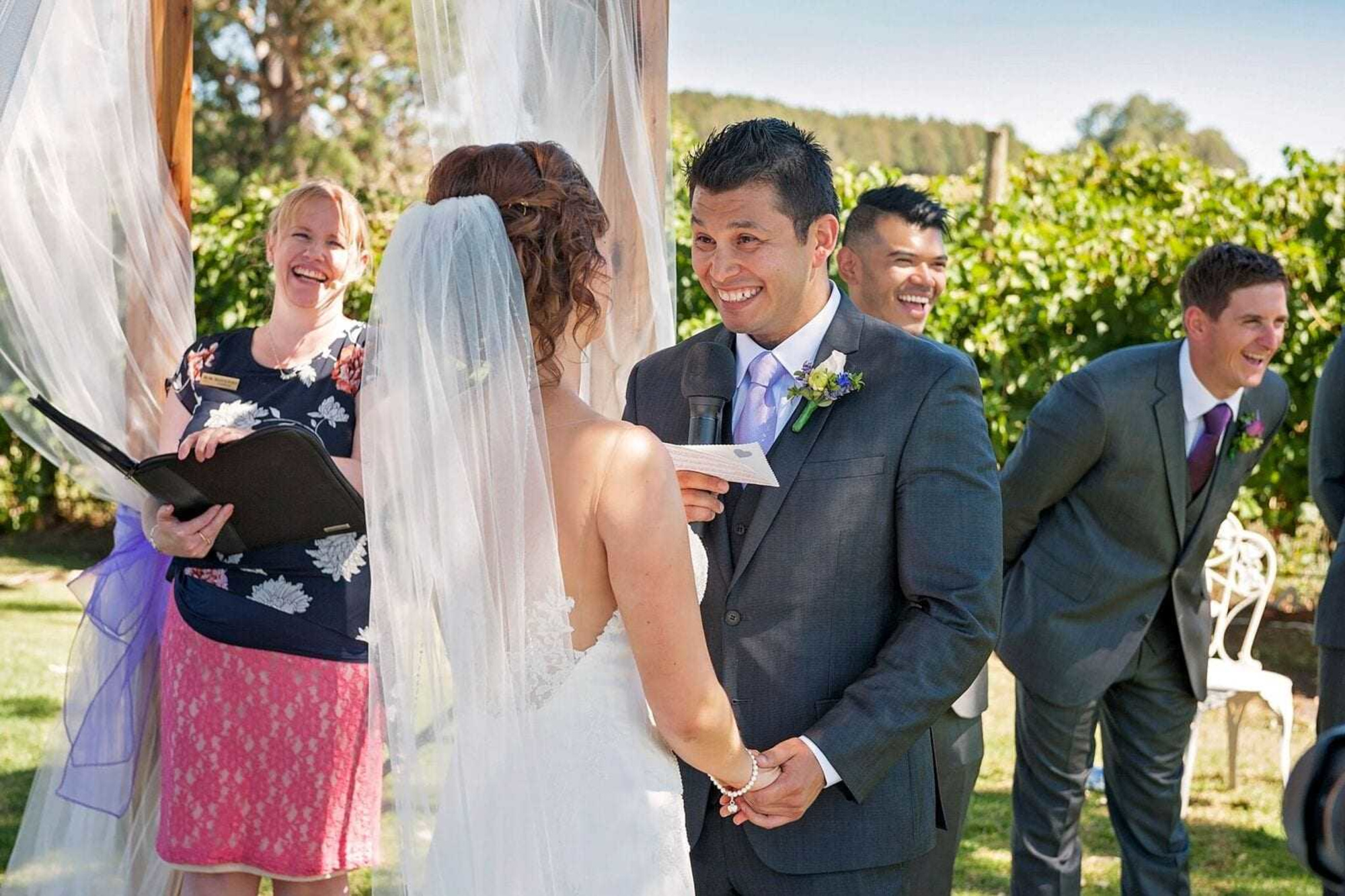 Bride and groom smiling and holding hands while exchanging vows during an outdoor wedding ceremony.