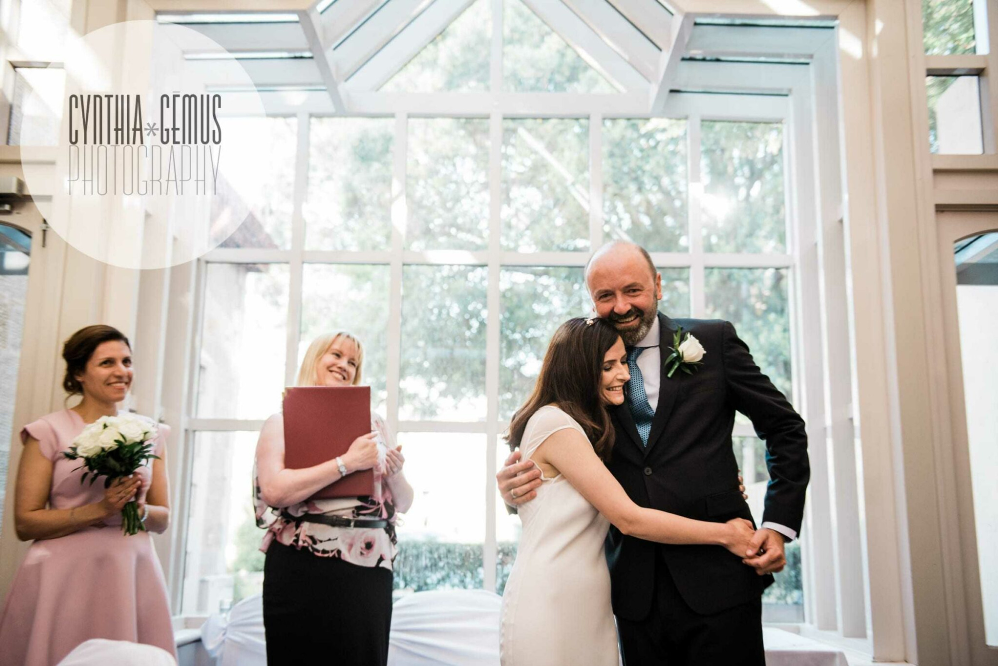 Joyful indoor wedding ceremony with the couple embracing under large bright windows.
