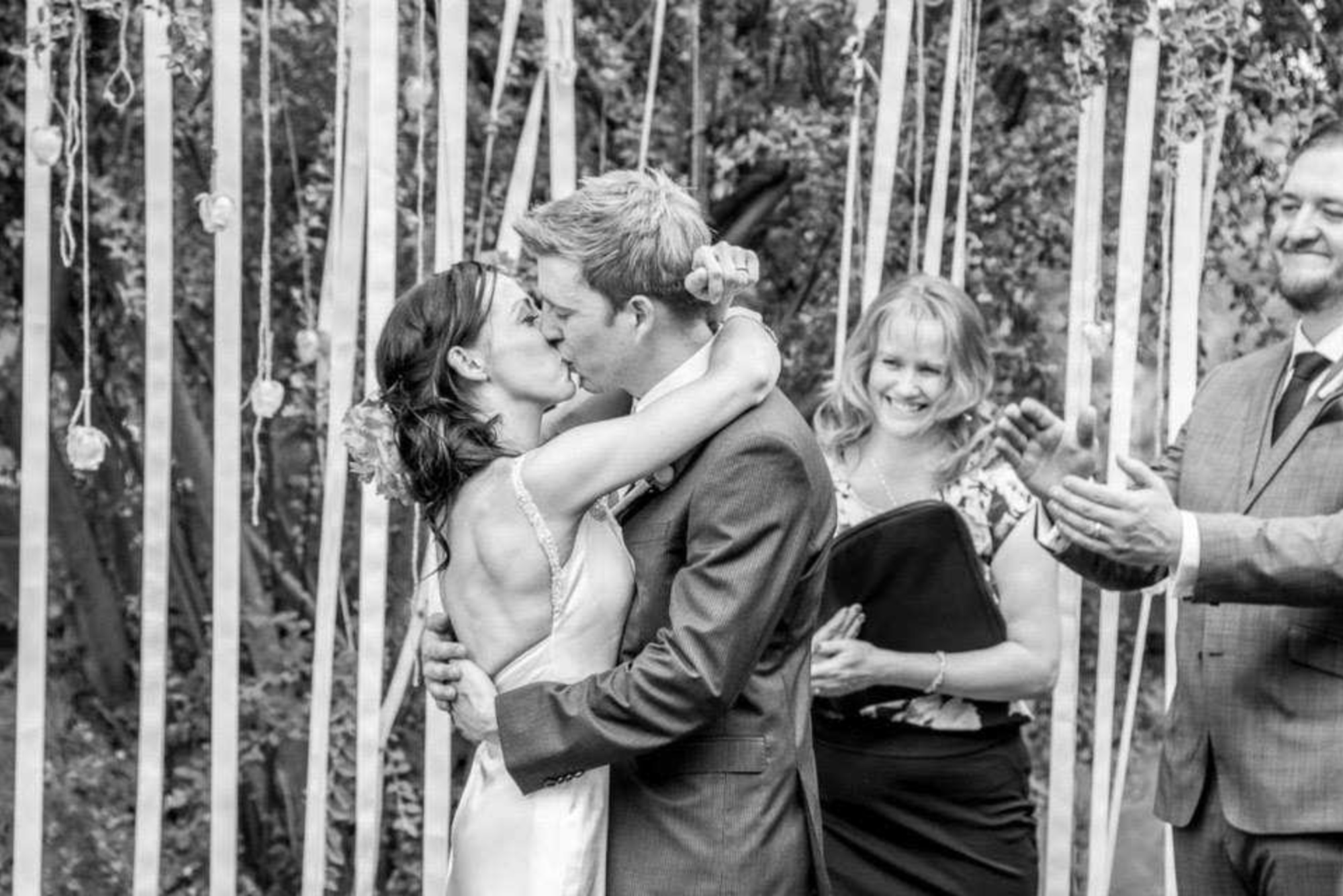 Bride and groom share a kiss during their outdoor wedding ceremony as guests smile and applaud.