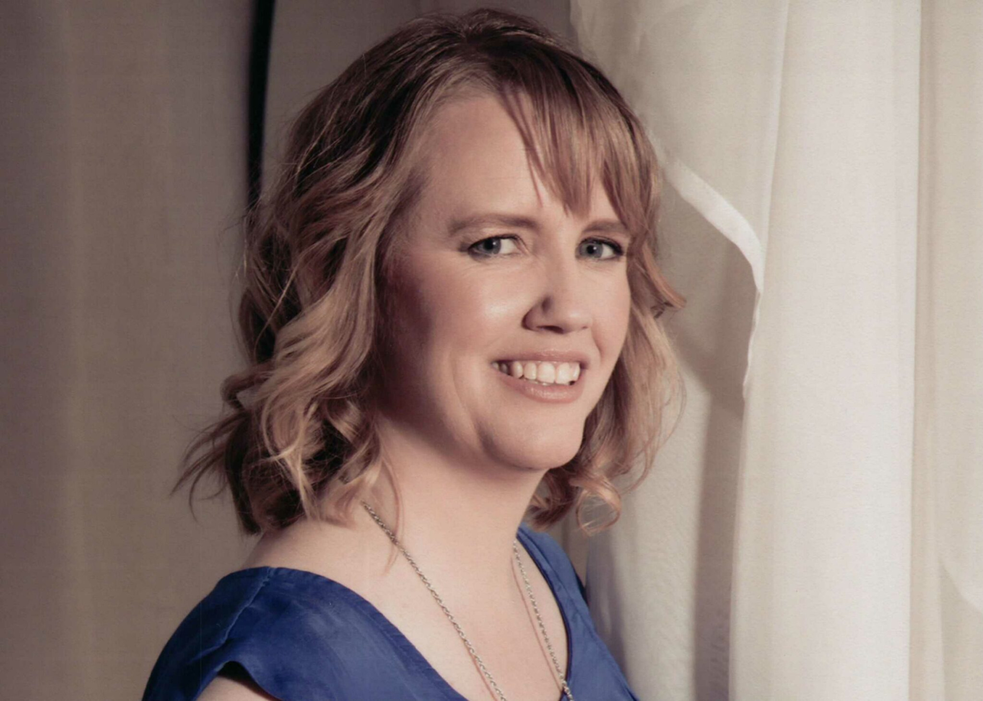 Professional headshot of a smiling woman in a blue dress standing beside light curtains.