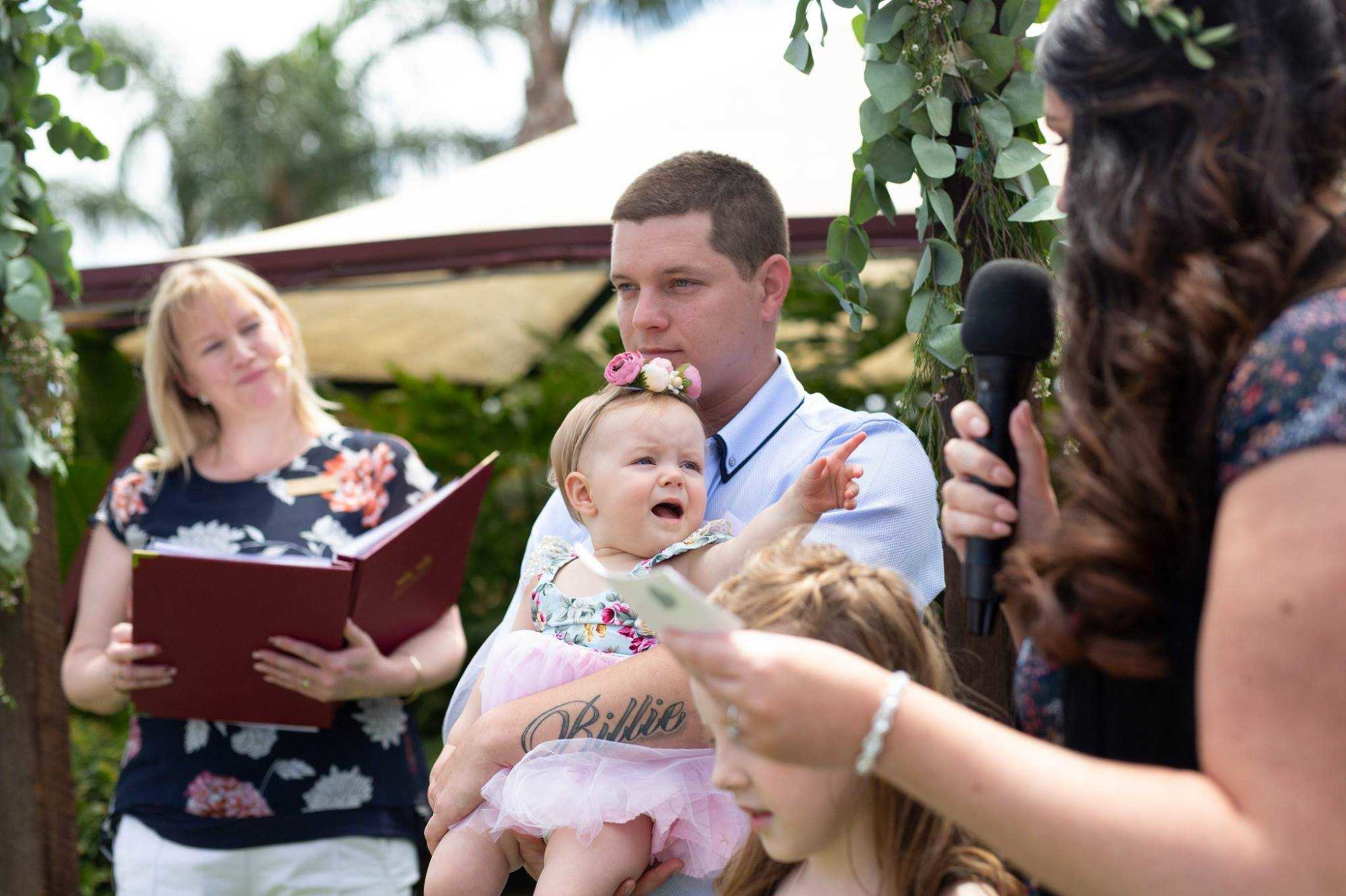 A man holding a baby during an outdoor wedding ceremony as a celebrant and guest speak nearby.