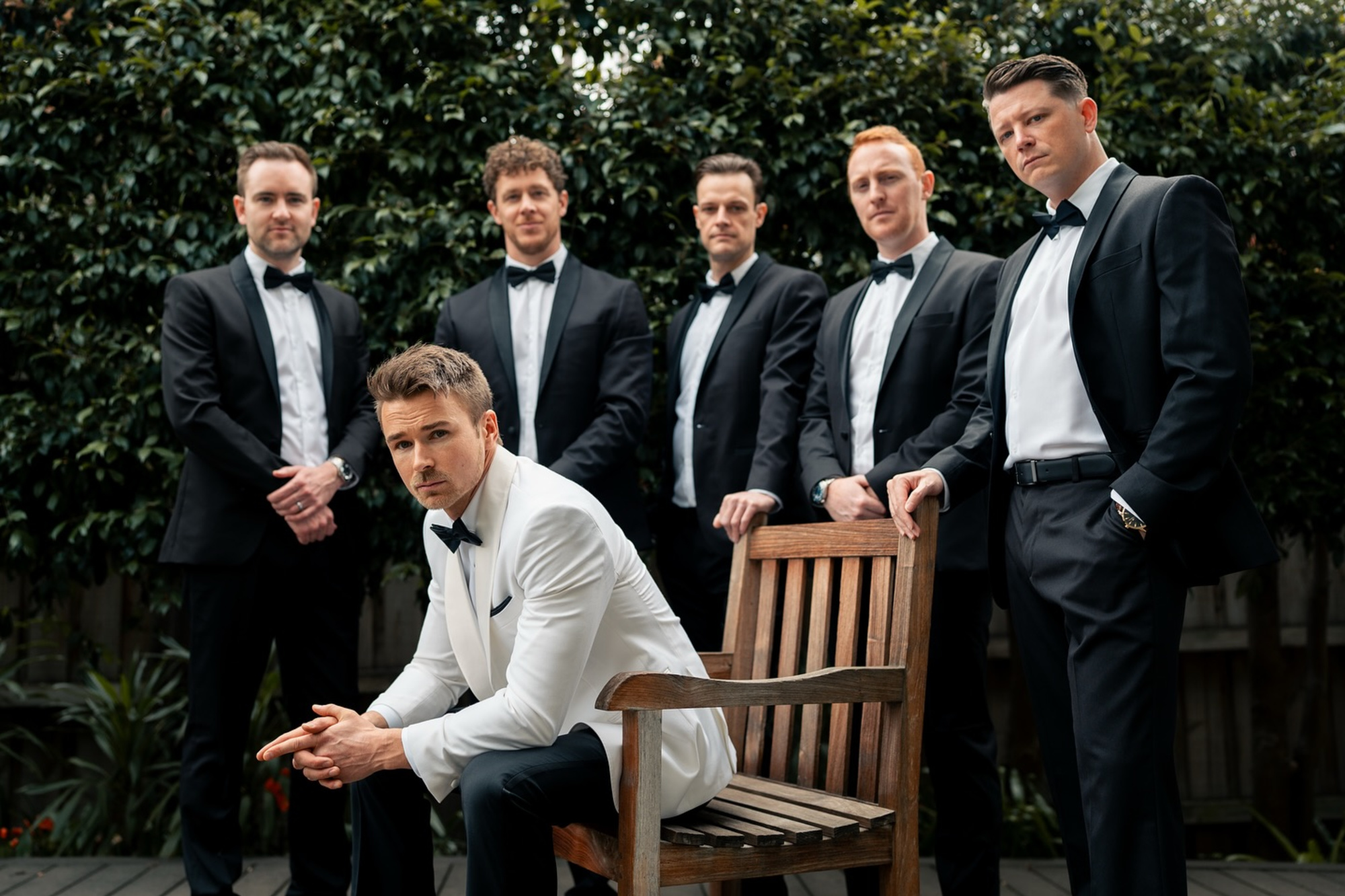 Groom in a white tuxedo jacket poses with his groomsmen in black suits in an outdoor setting.