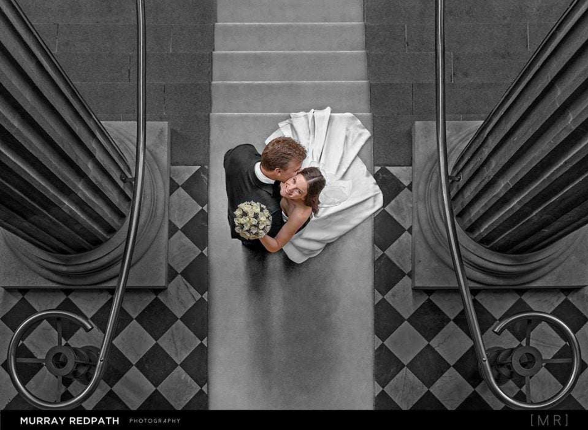 Overhead view of a bride and groom embracing on a grand staircase framed by tall columns and checkered floor tiles.