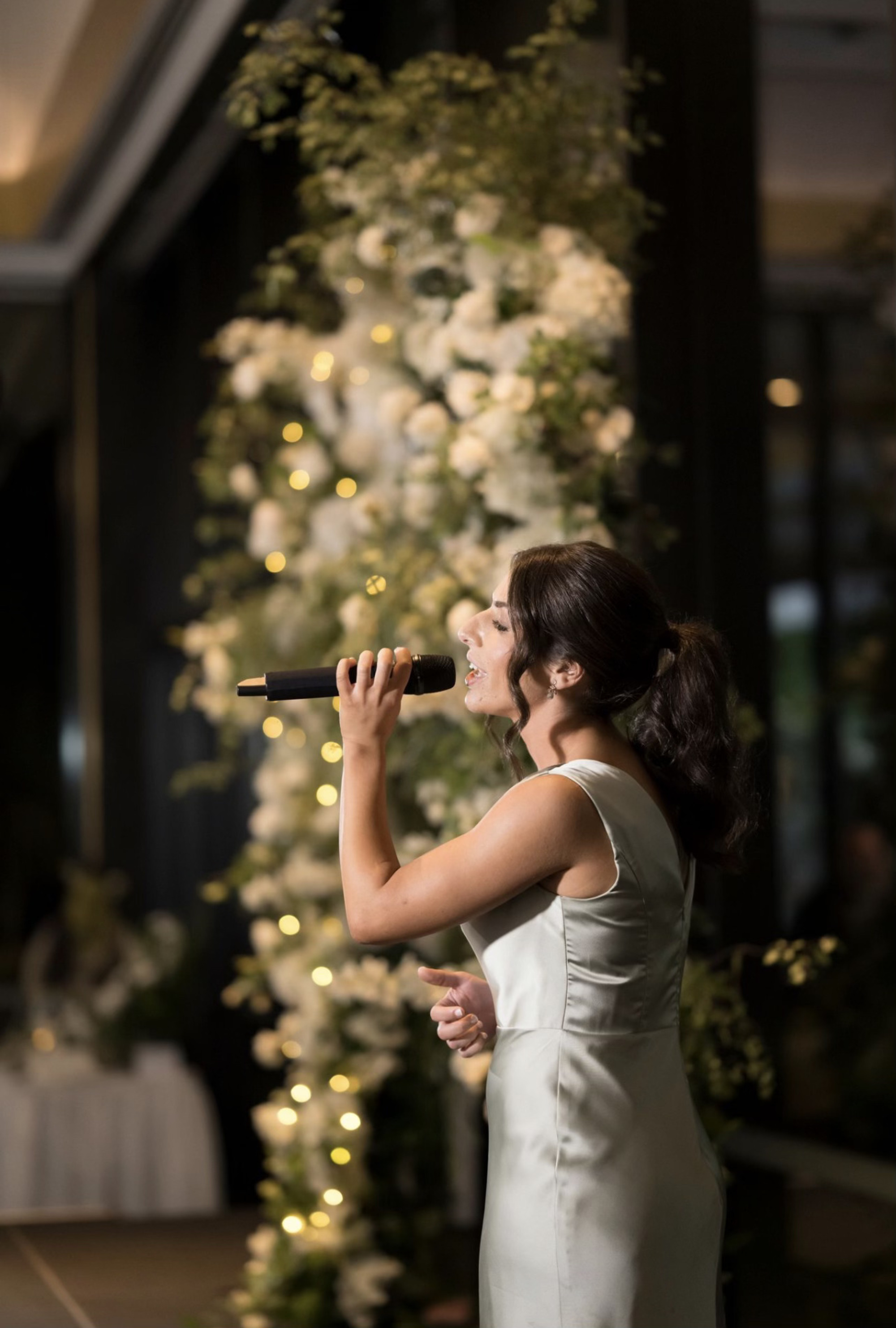 Female singer performs at a wedding in front of a floral arch with warm fairy lights.