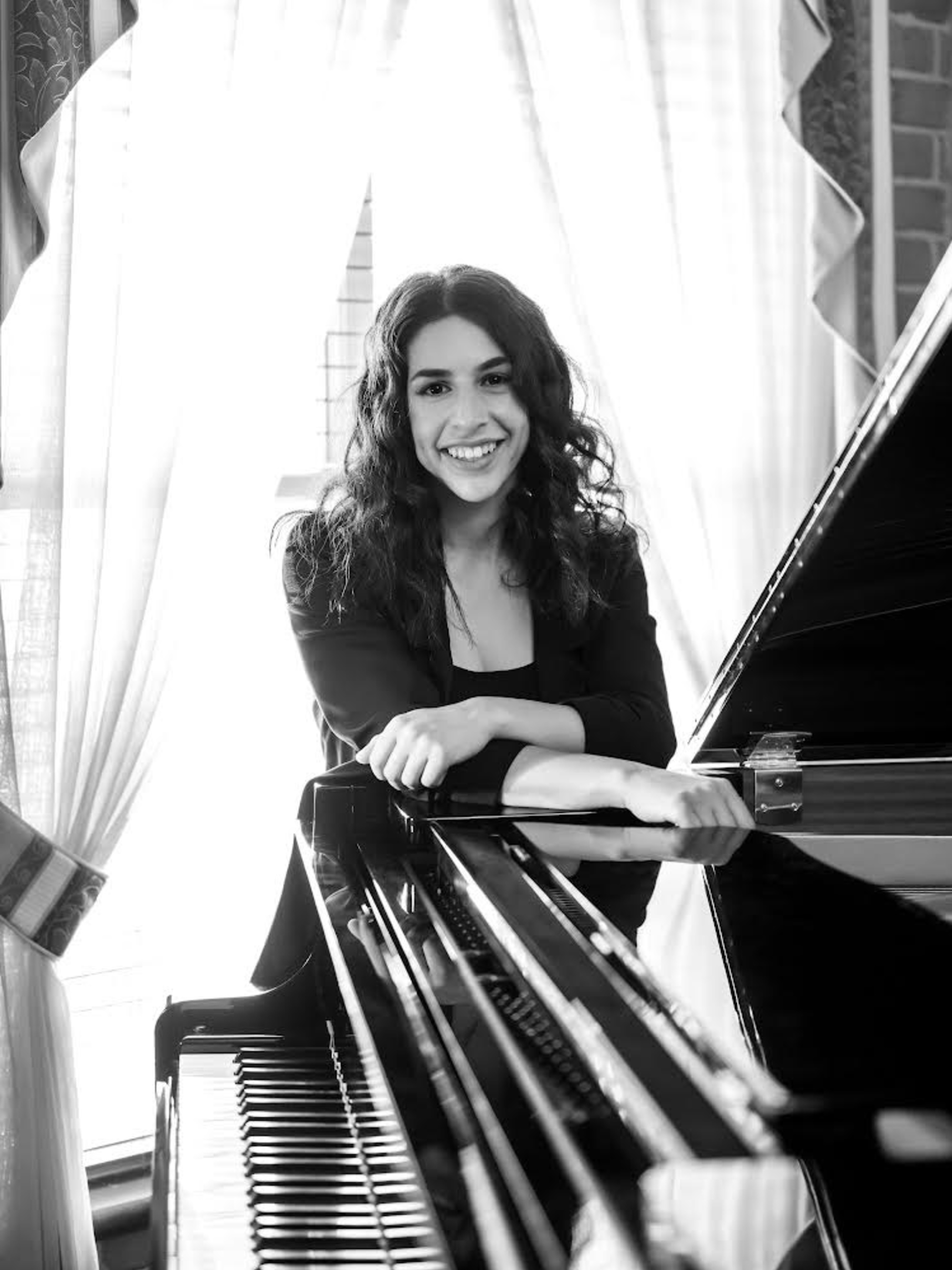 Smiling pianist leaning on a grand piano in front of bright window curtains.