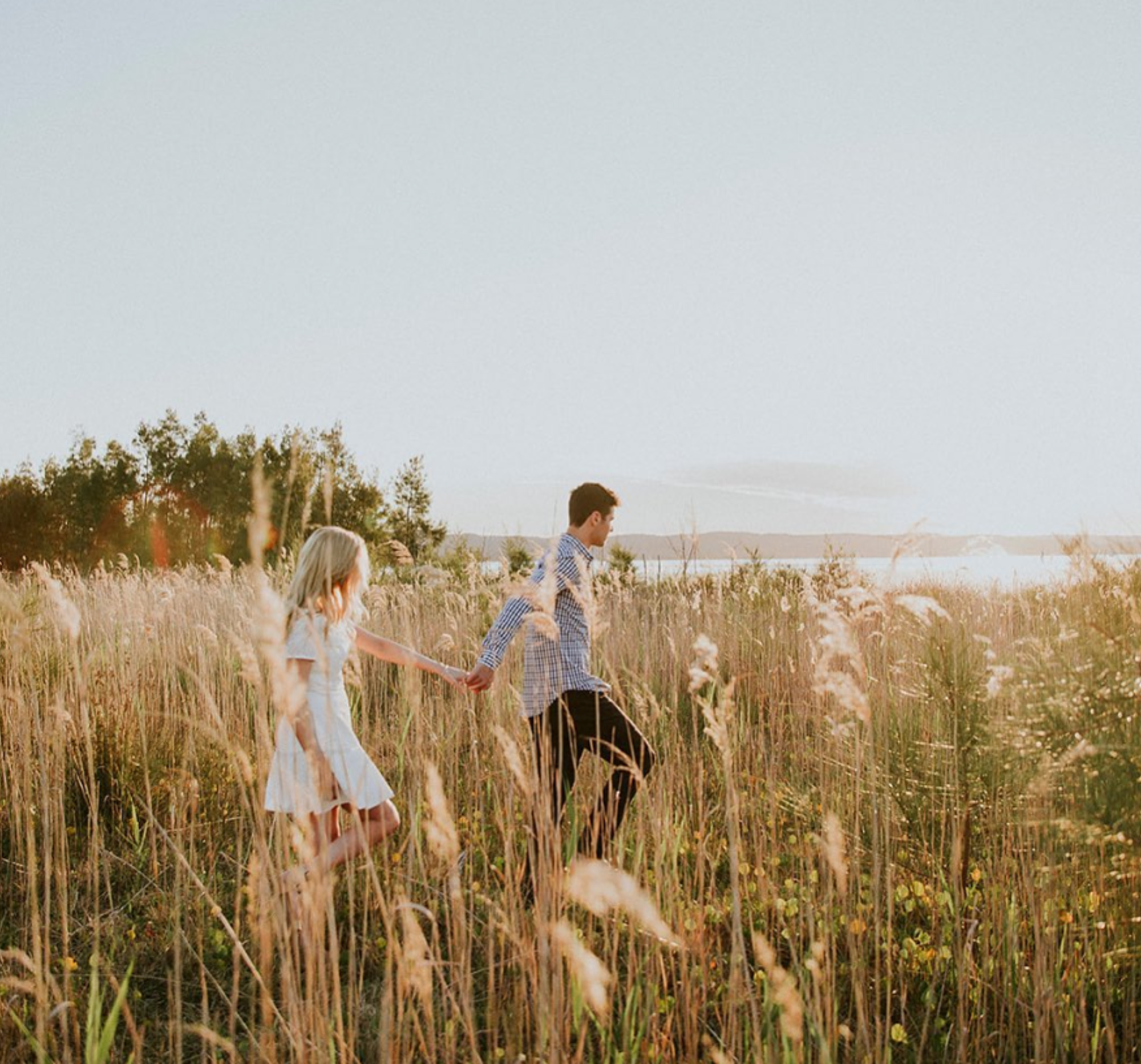 A couple holds hands while walking through a tall grassy field at sunset near the water.