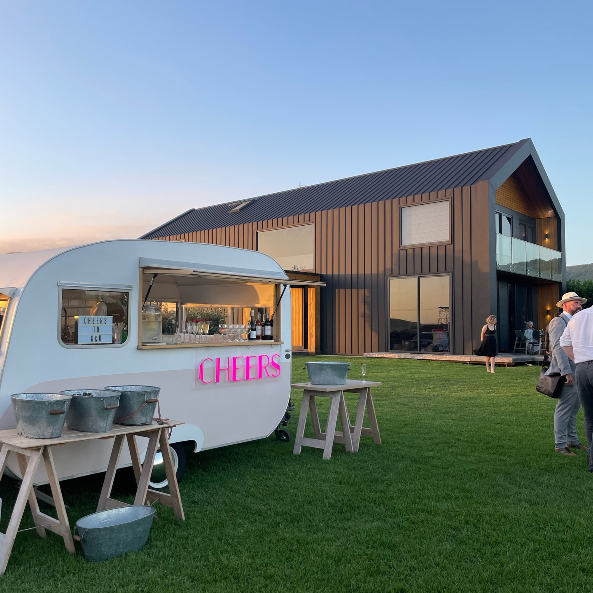 Mobile bar trailer serving drinks on a lawn outside a modern barn-style wedding venue at sunset.