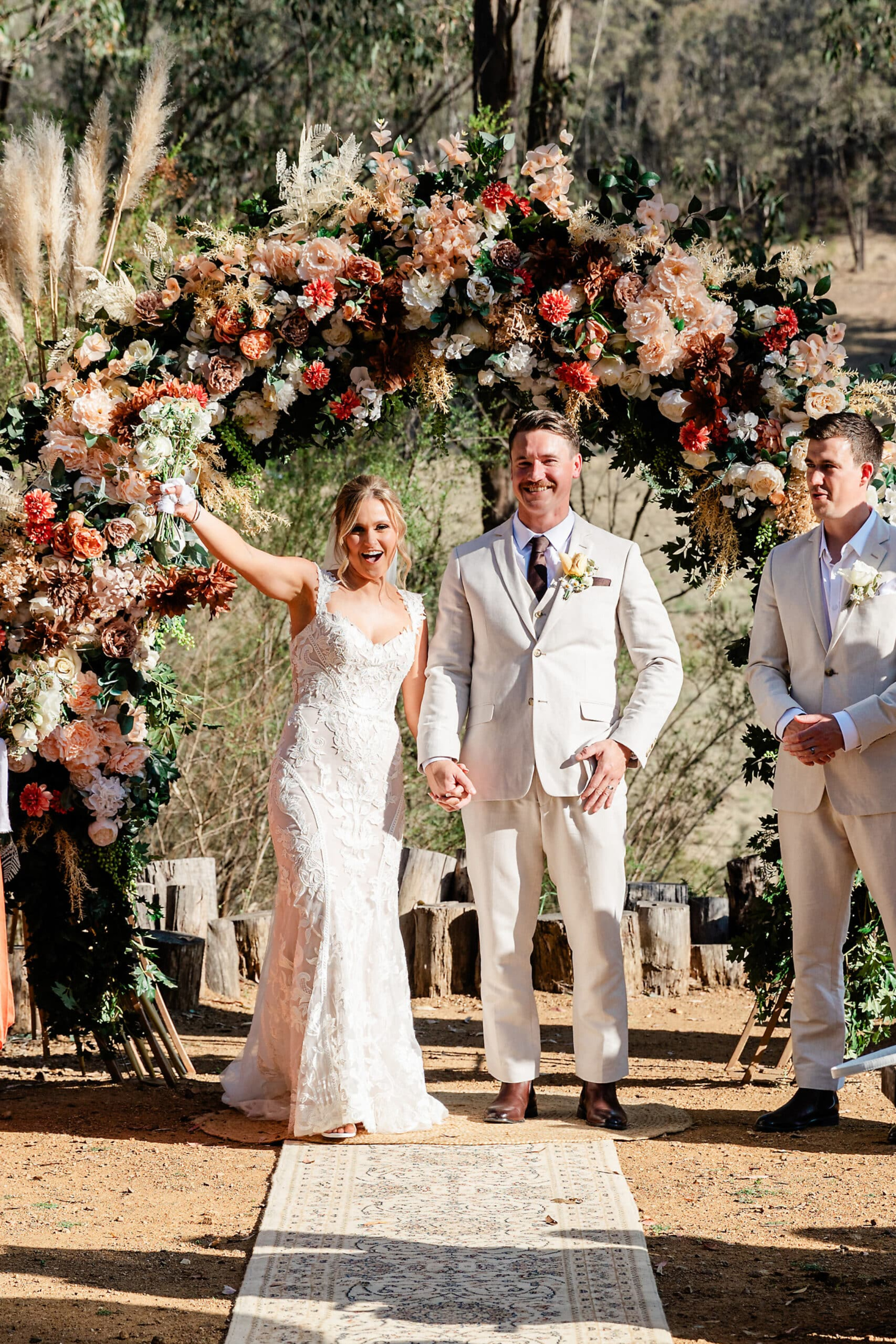 Bride and groom celebrate under a lush floral ceremony arch at an outdoor rustic wedding.