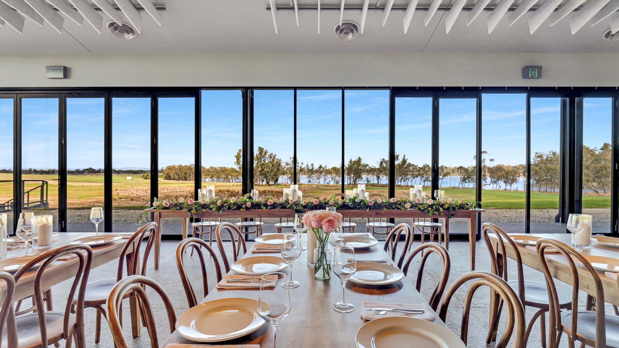Elegant wedding reception tables with pink roses and candle decor overlooking a scenic lakeside landscape through large windows.