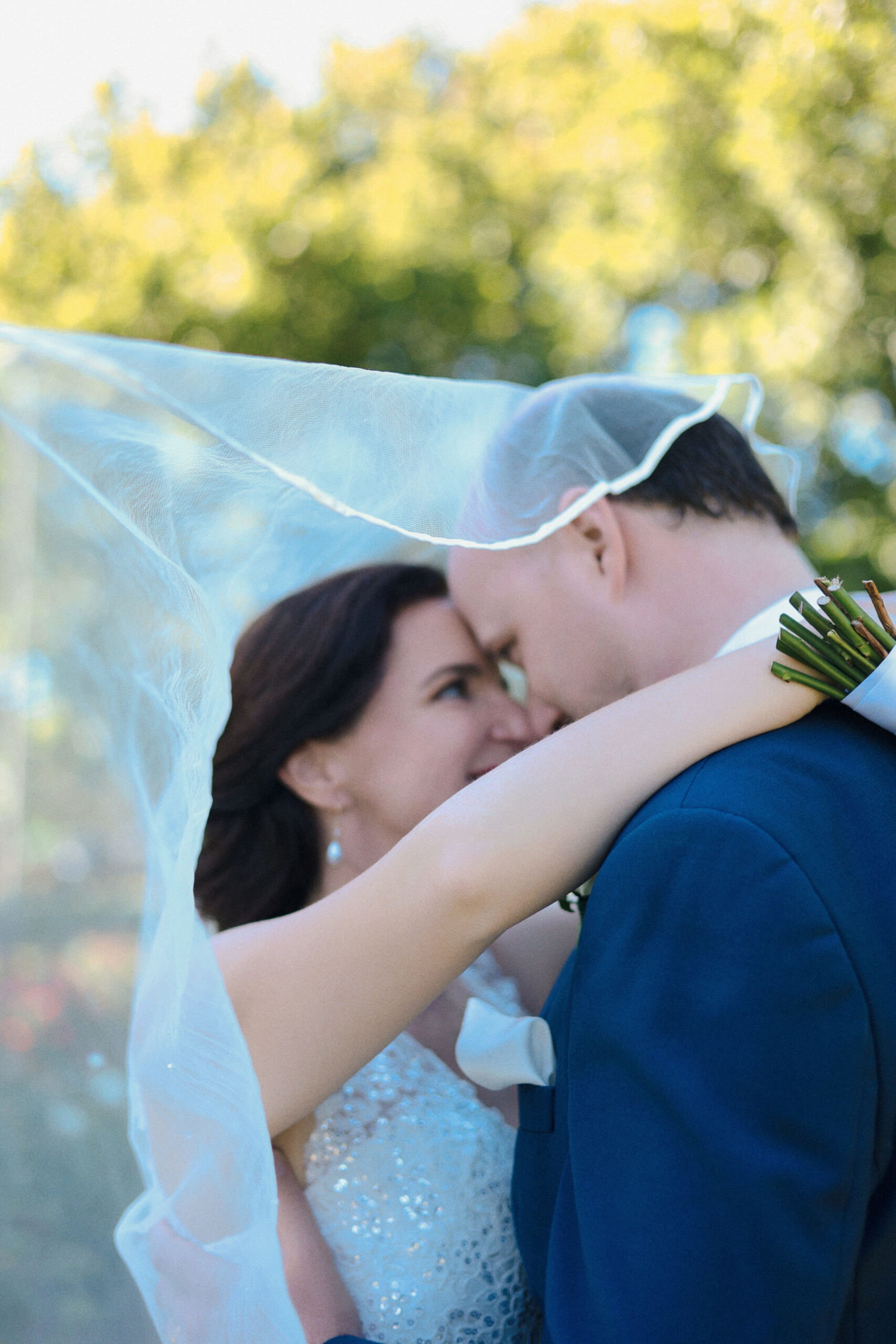 Bride and groom embrace under a flowing veil during an outdoor wedding portrait.