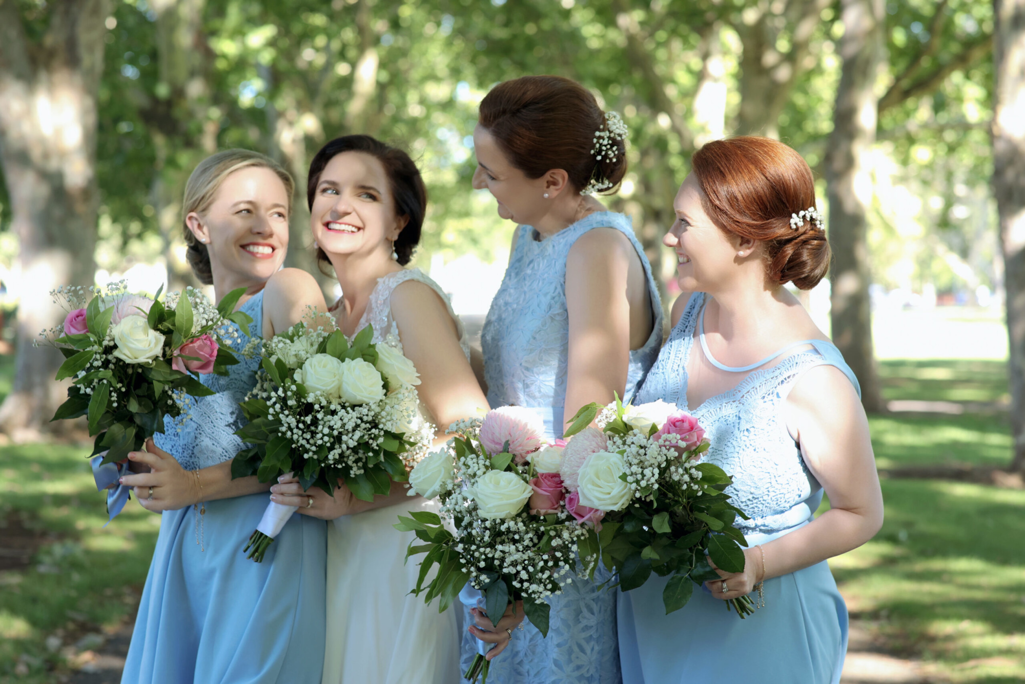 Bride and bridesmaids in light blue dresses holding pastel flower bouquets in a sunny garden.