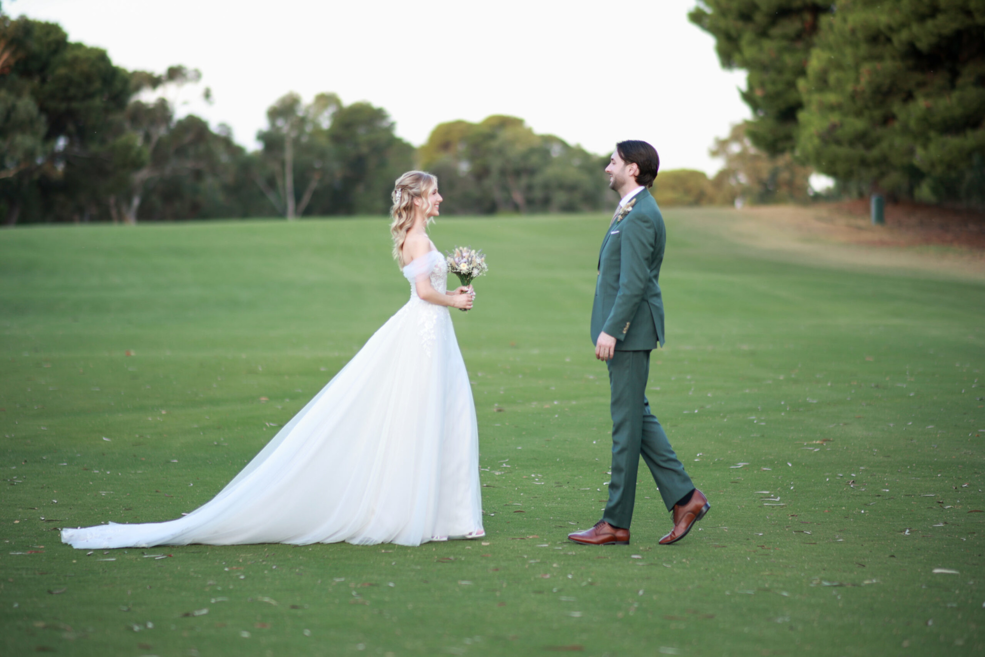 Bride and groom share a first look on a wide green lawn surrounded by trees.