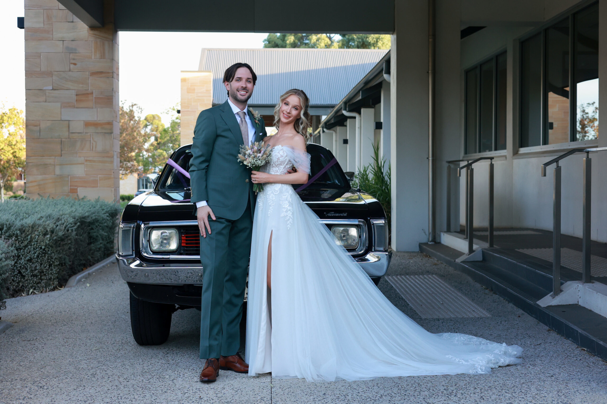 Bride and groom pose in front of a vintage black car outside a modern wedding venue.