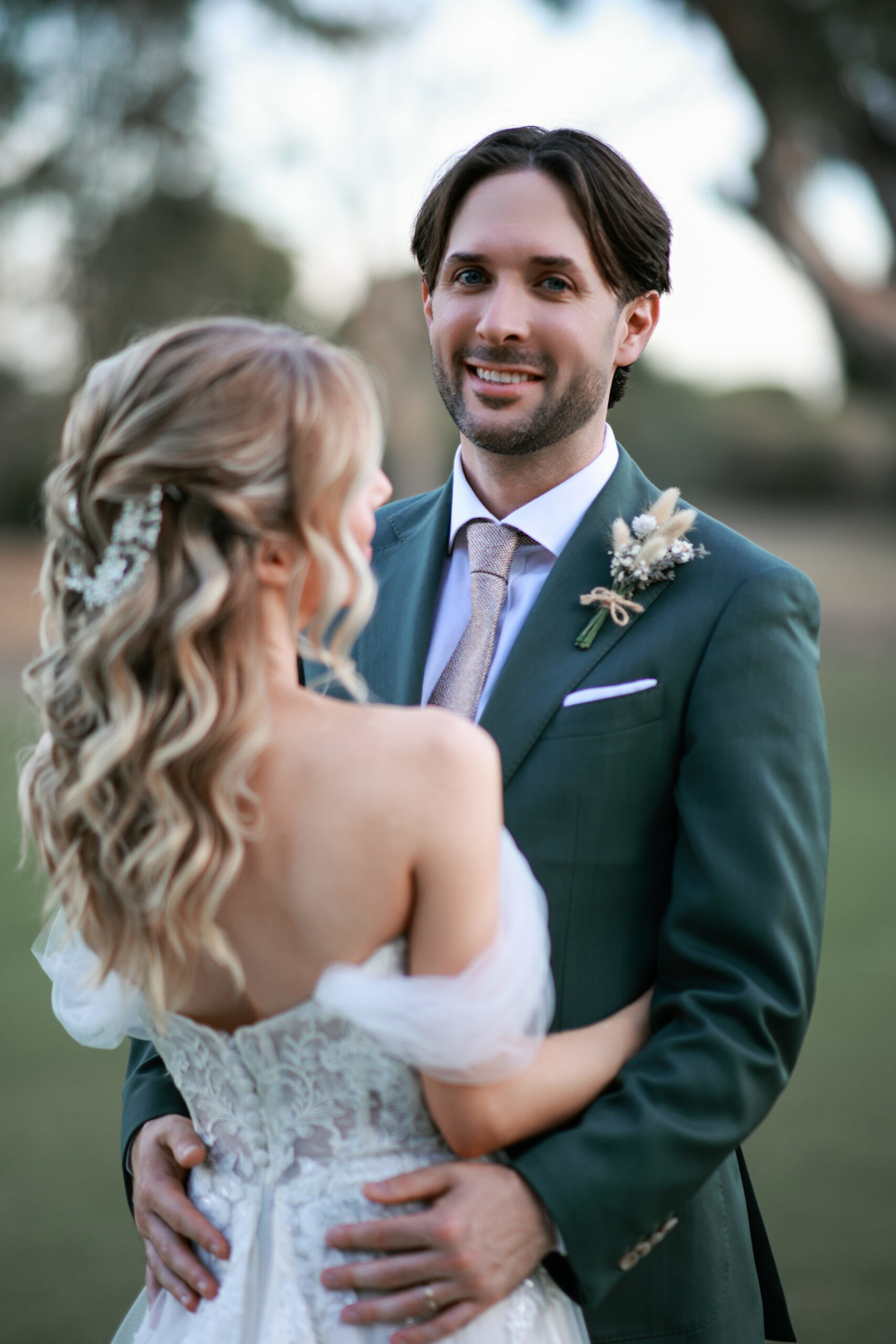 Outdoor wedding portrait of a smiling groom in a green suit holding his bride in a lace off-shoulder gown.
