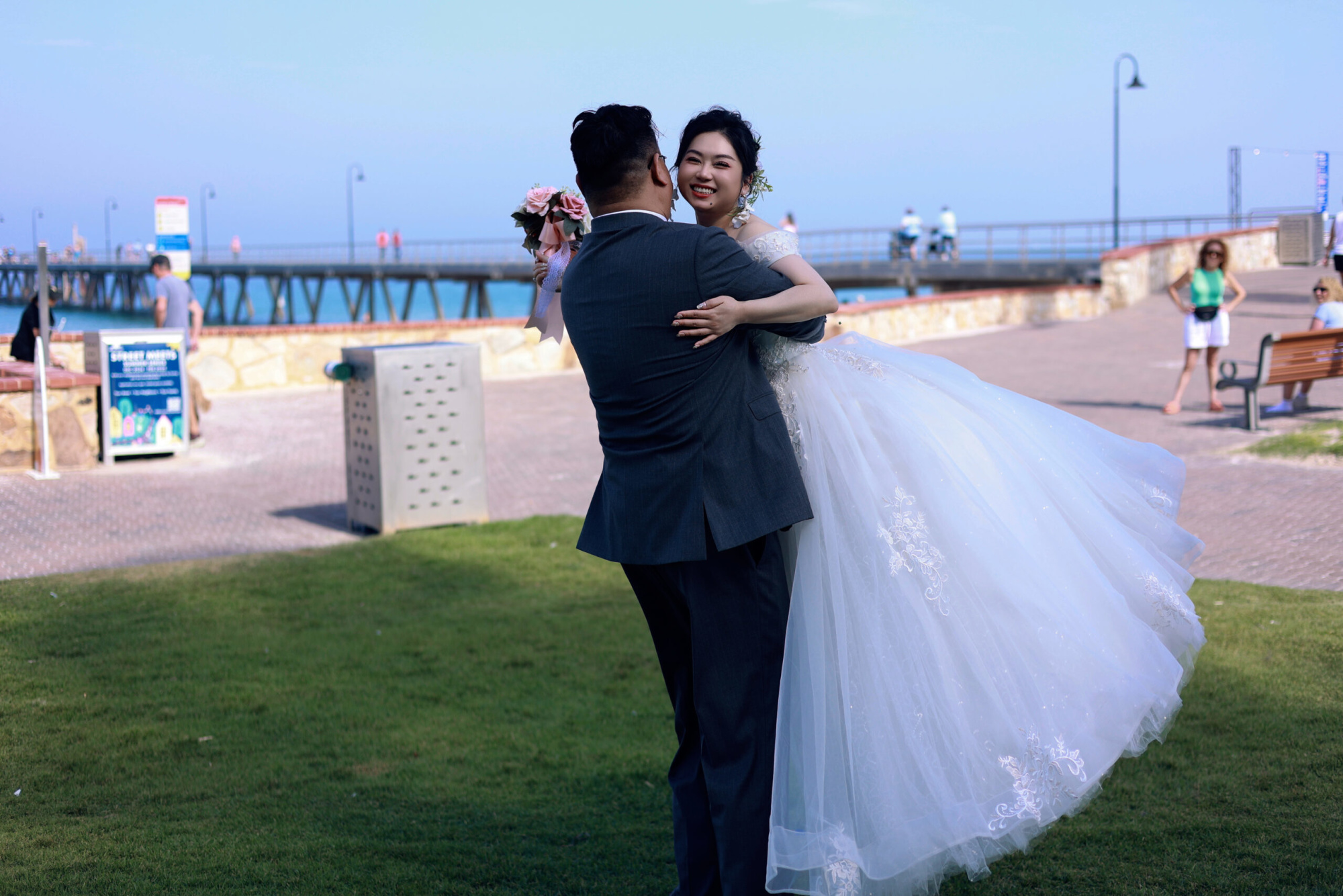 Groom carries smiling bride in a flowing white gown by a seaside pier on a sunny day.