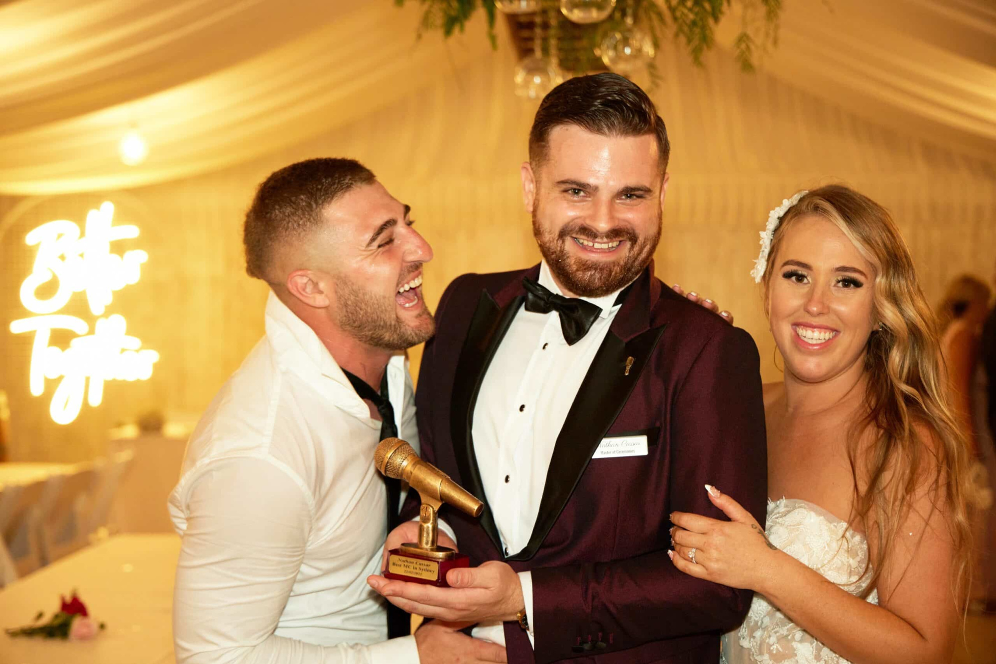Bride and groom laugh with their wedding MC holding a gold microphone award in a warm-lit reception tent.