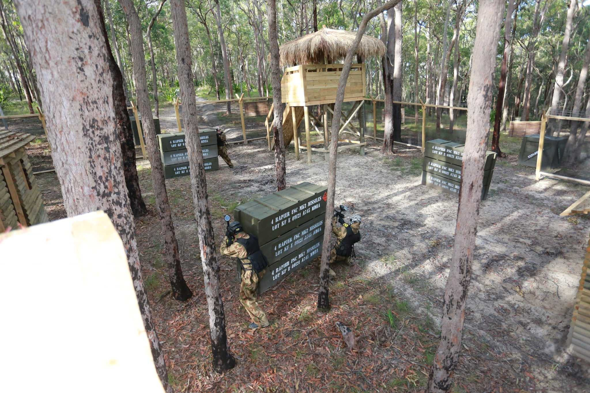 People in camouflage play paintball in a wooded outdoor arena with bunkers and a raised hut.