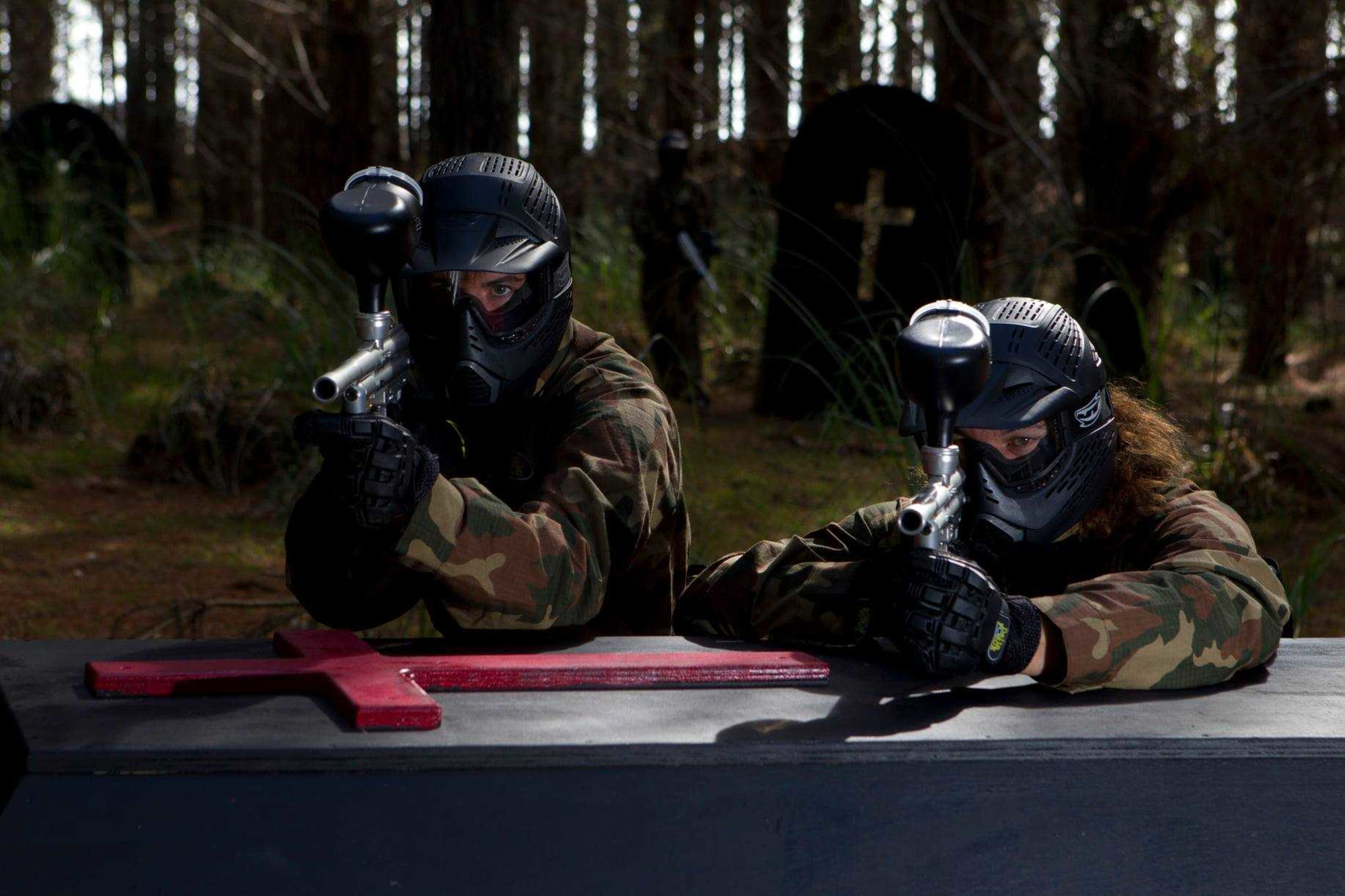 Two people in camouflage gear and masks aim paintball guns behind a barricade in a wooded field.
