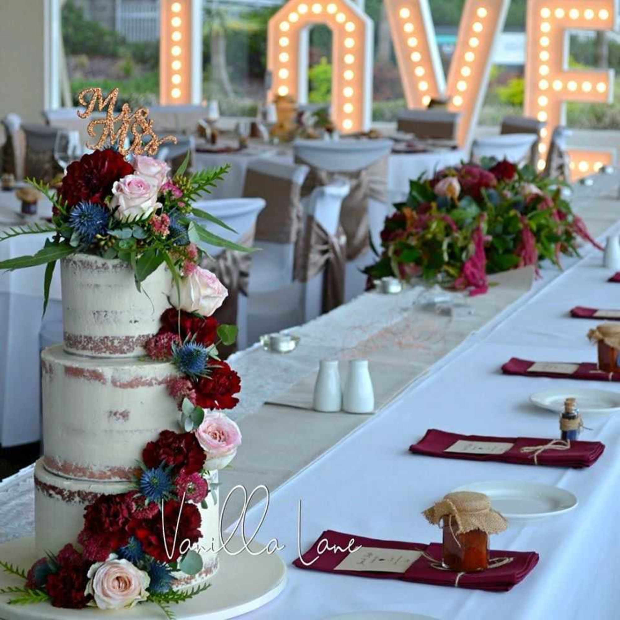 Rustic wedding reception table with floral naked cake and large illuminated LOVE sign in the background.