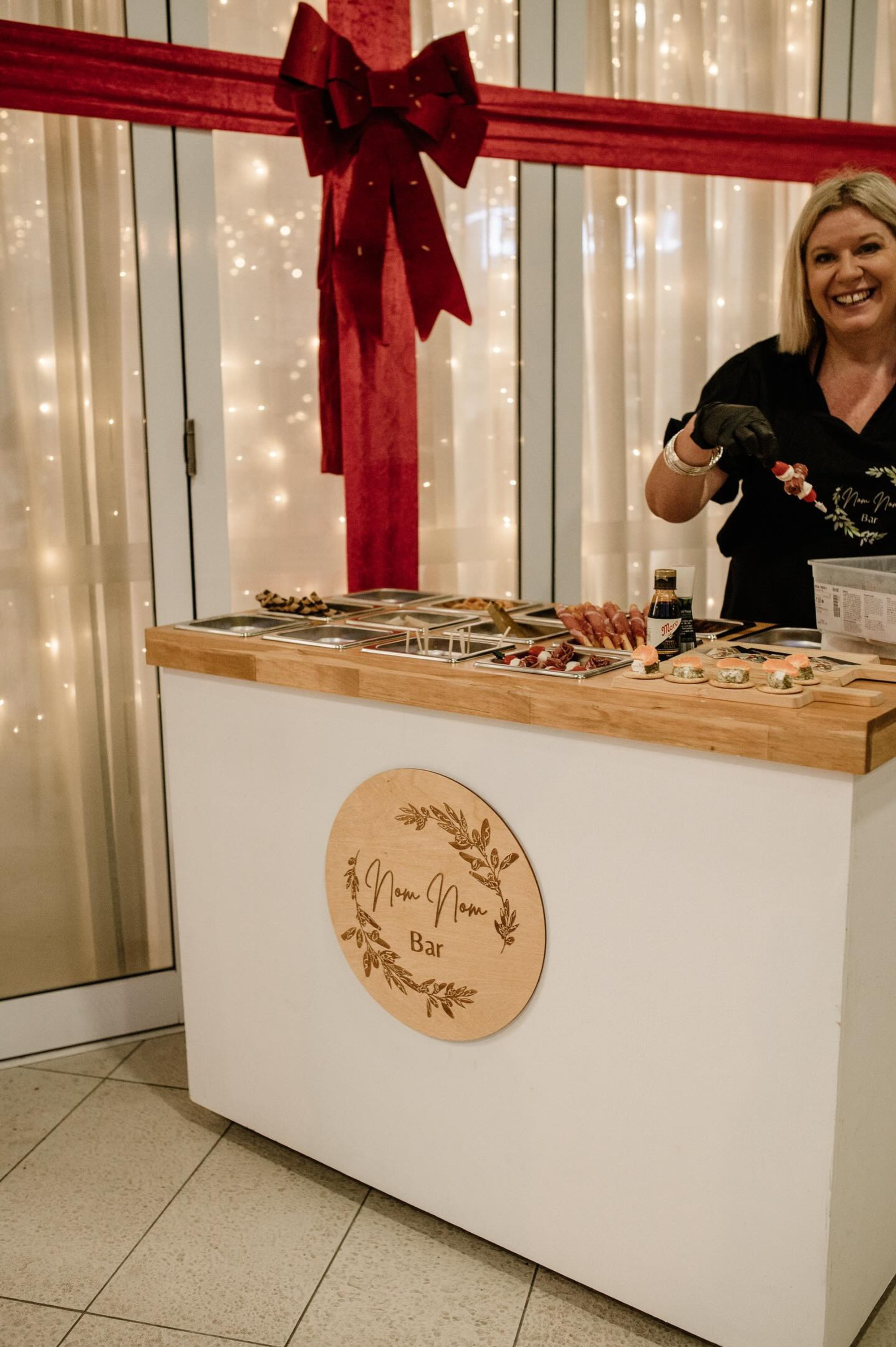 Caterer serving canapés from a stylish food bar in front of twinkling lights and a large red ribbon backdrop.