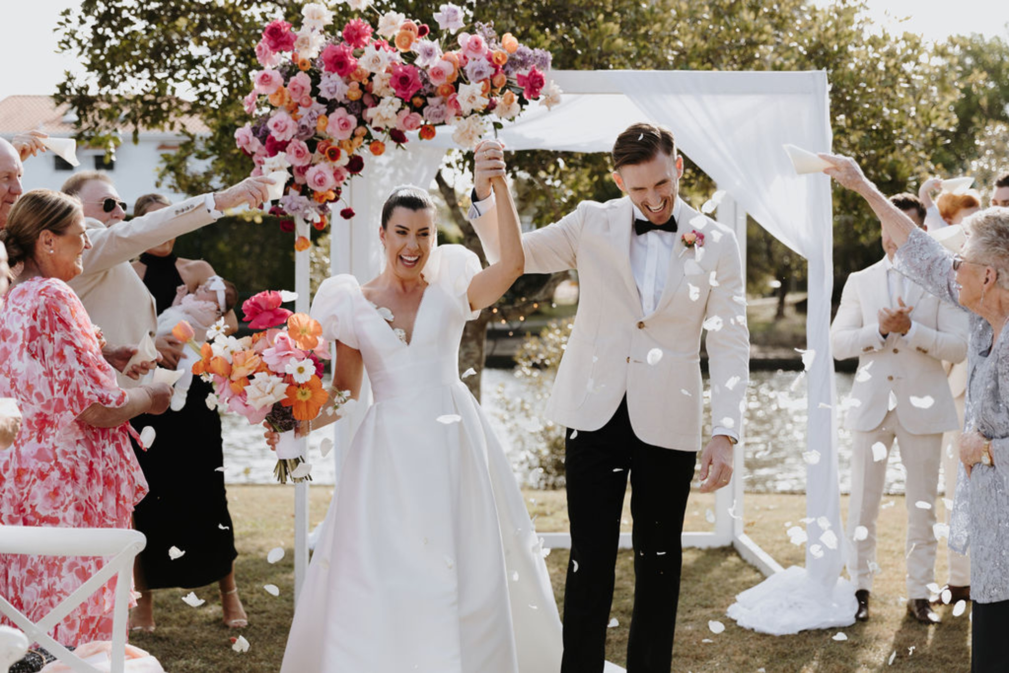 Joyful couple walk back down the aisle under a colorful floral arch as guests toss petals at their outdoor lakeside wedding.
