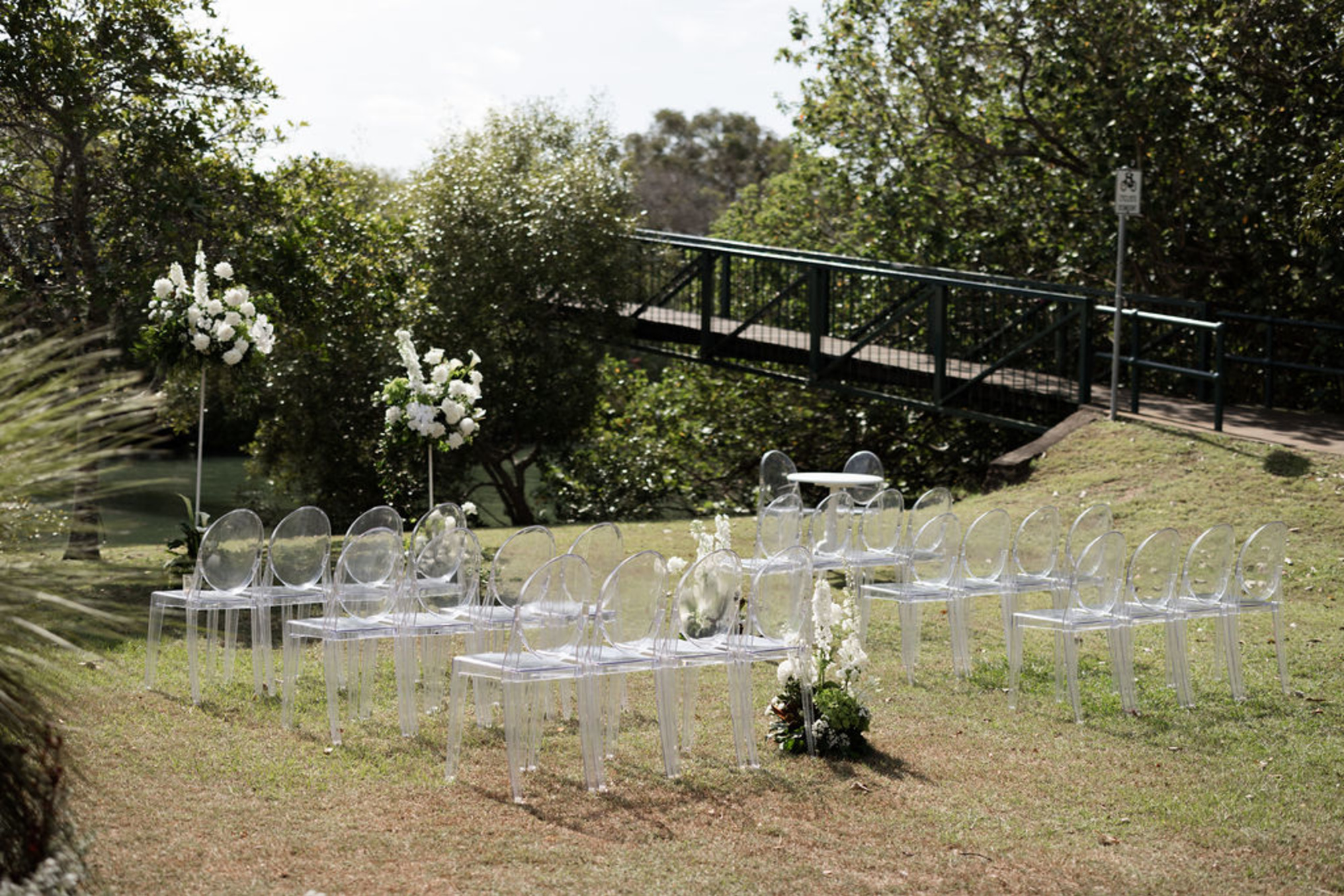 Modern outdoor wedding ceremony setup with clear chairs and white floral arrangements by a garden bridge.