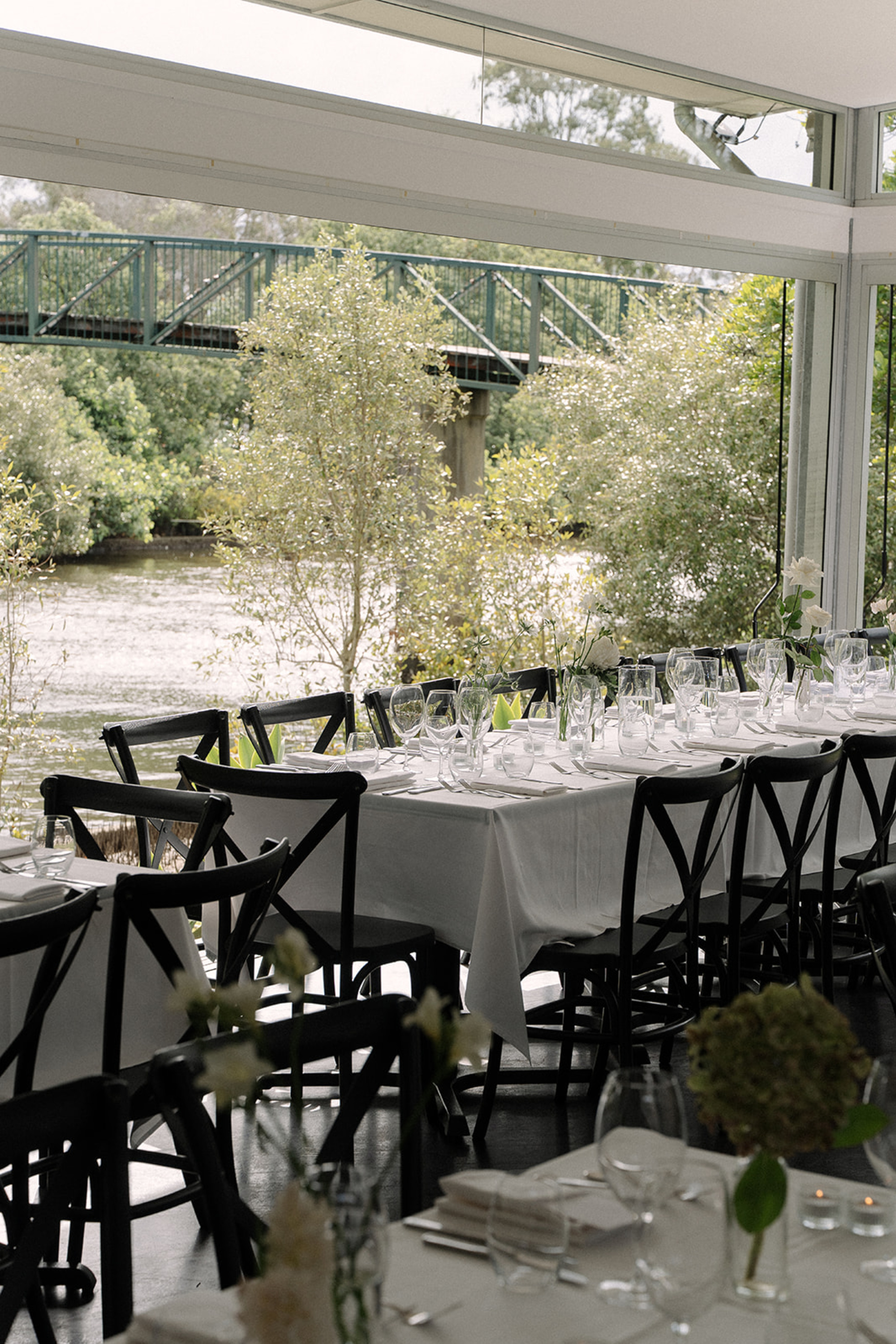 Elegant riverfront wedding reception tables set with white linens and glassware beside large windows overlooking trees and a bridge.