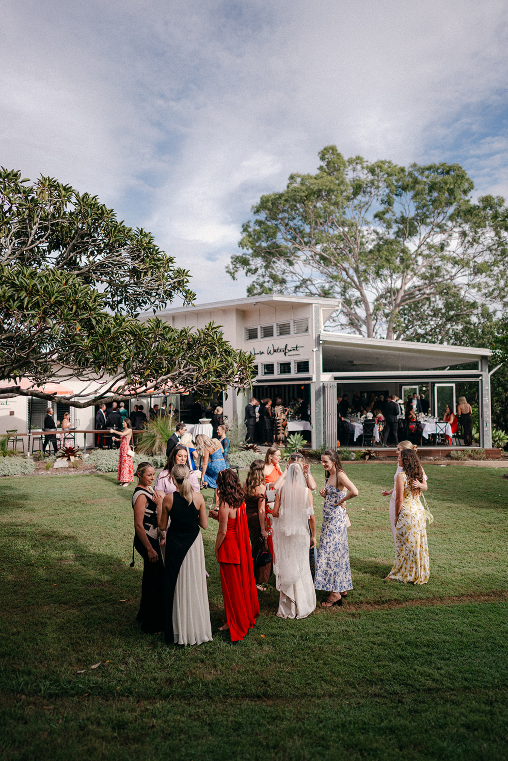 Wedding guests mingle on a grassy lawn outside a modern waterfront venue surrounded by trees.