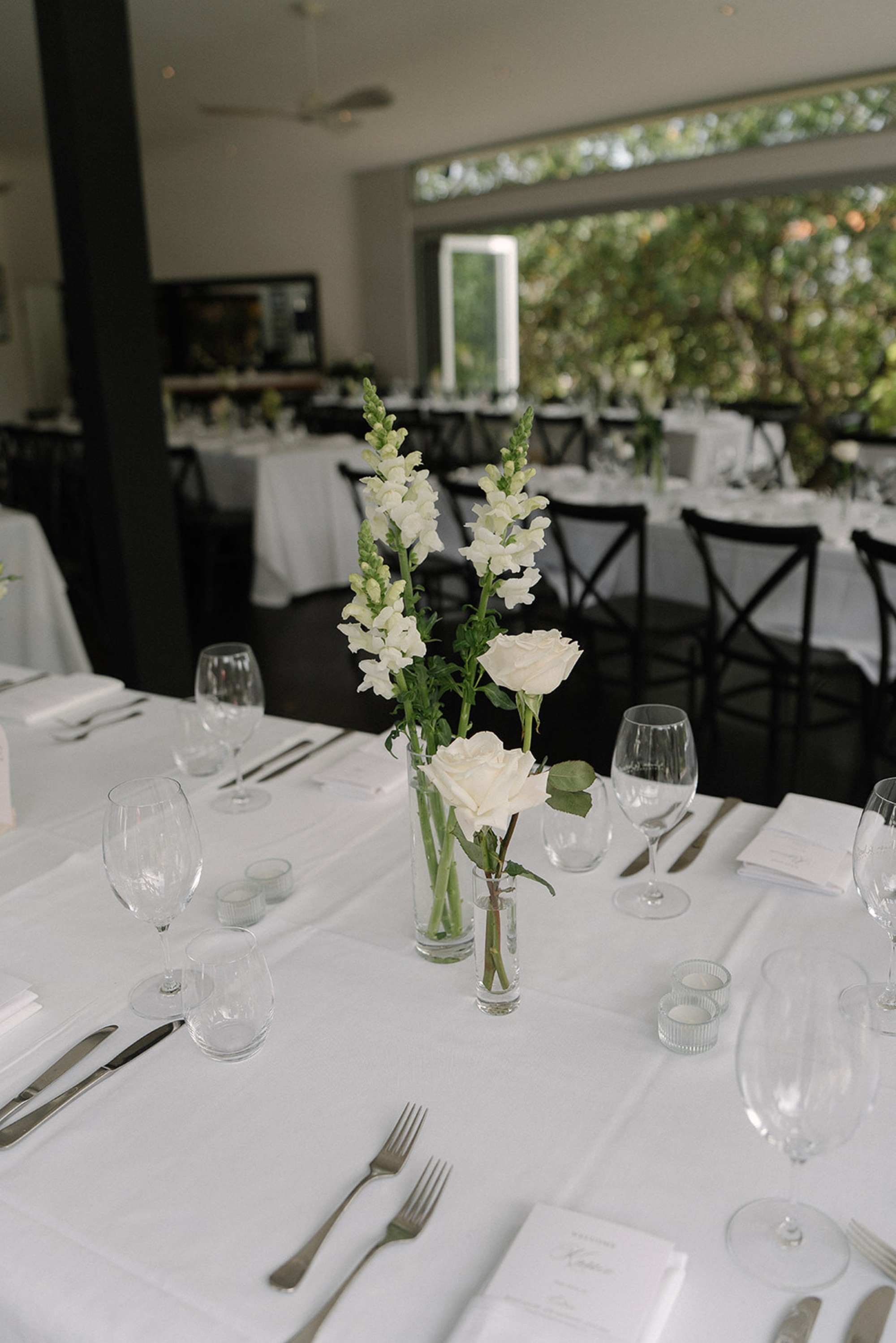Elegant wedding reception table with white floral centerpieces, glassware, and simple place settings in a bright indoor venue.