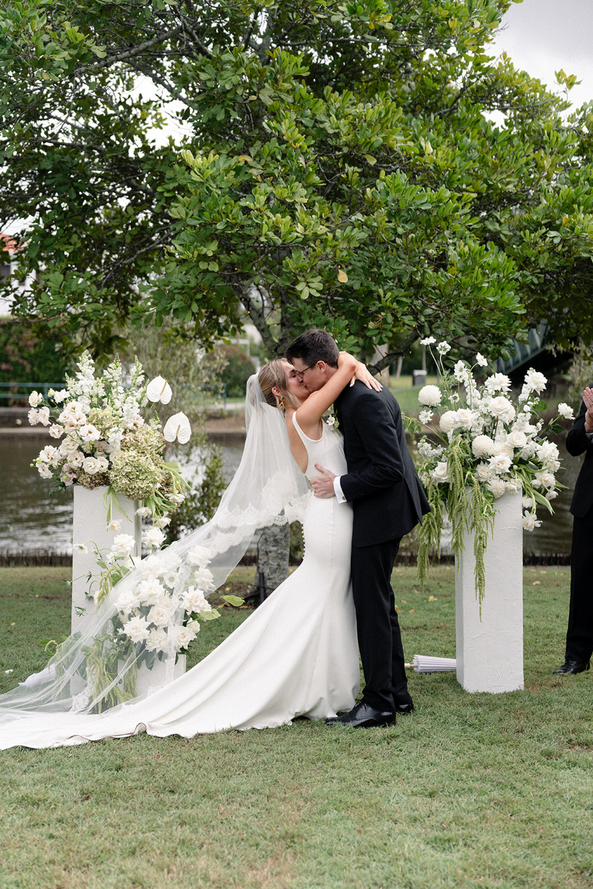 Bride and groom share a kiss at an outdoor ceremony framed by white floral arrangements and greenery.