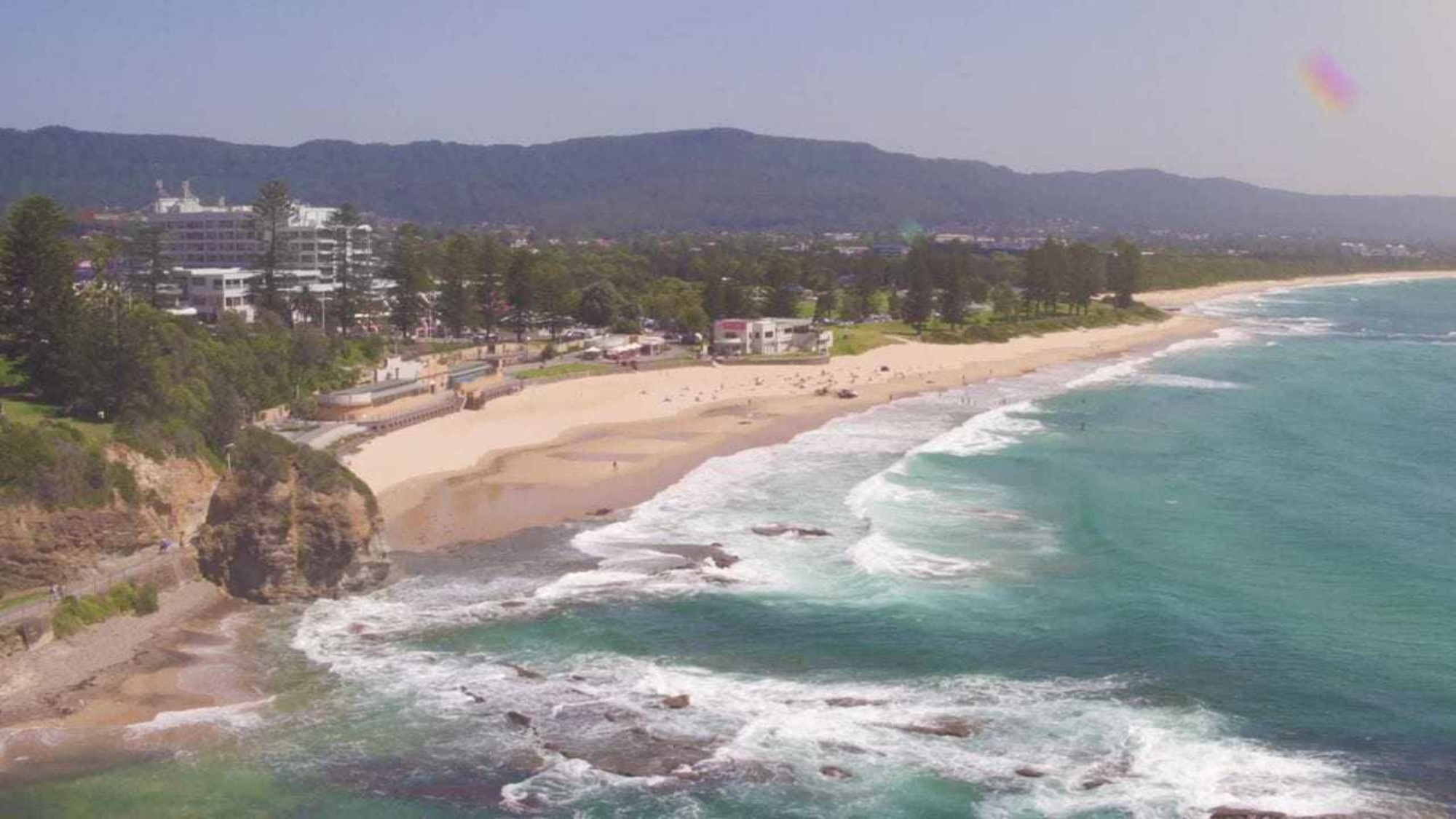 Aerial view of a scenic beach with turquoise waves, sandy shore, and nearby coastal buildings and greenery.
