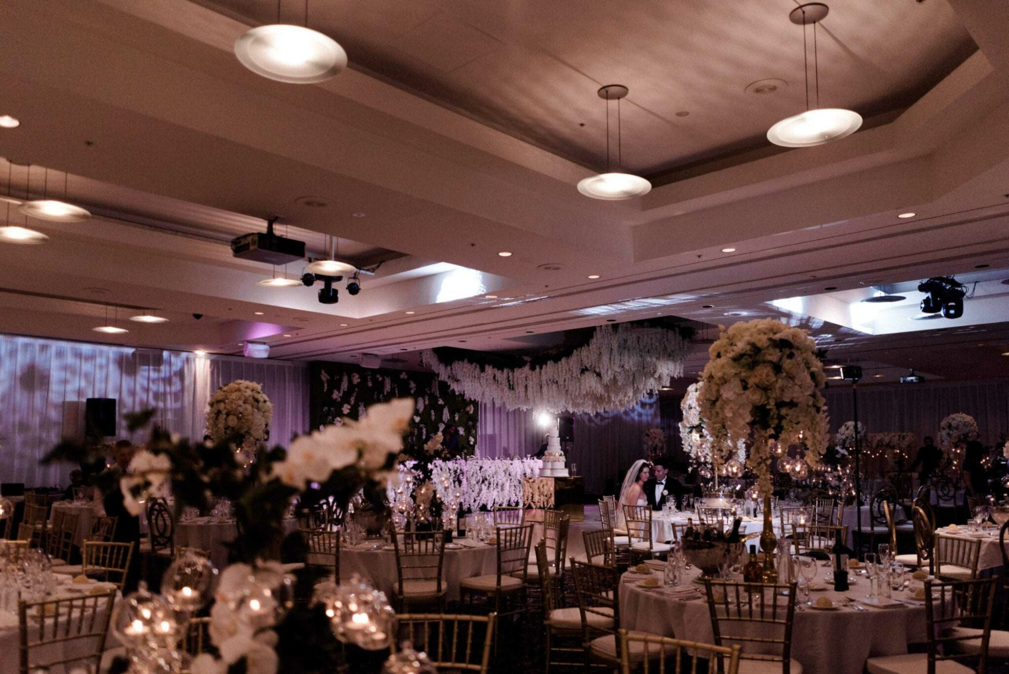 Luxurious indoor wedding reception with tall white floral centerpieces, gold chairs, and soft romantic lighting.