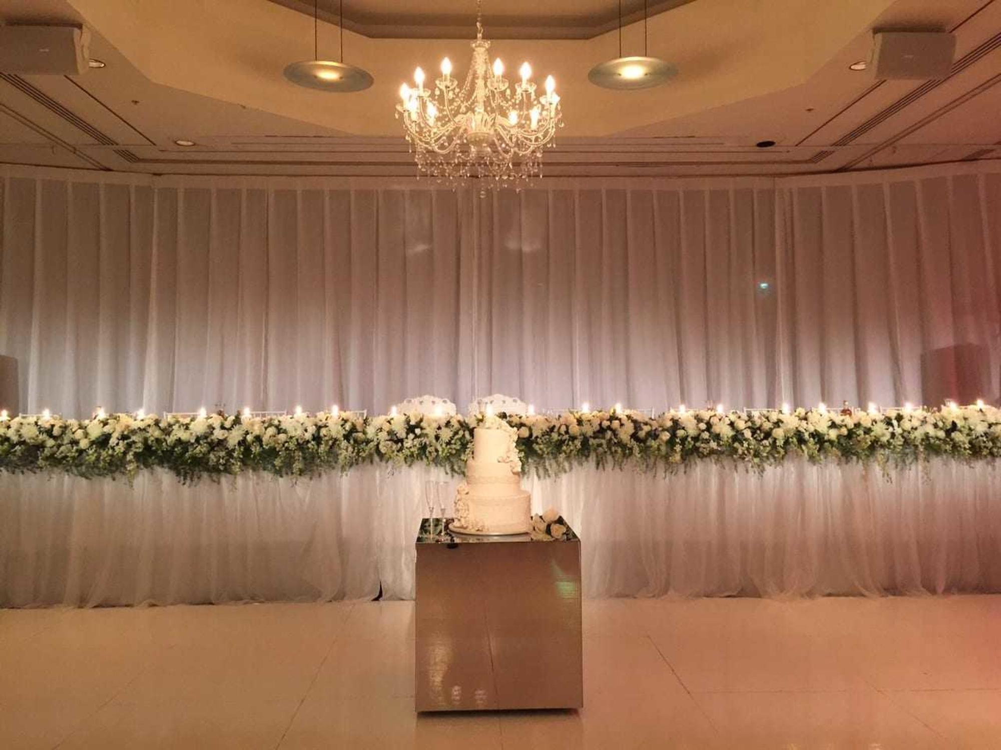 Elegant wedding reception setup with a tiered white cake, long floral arrangement, and chandelier lighting.