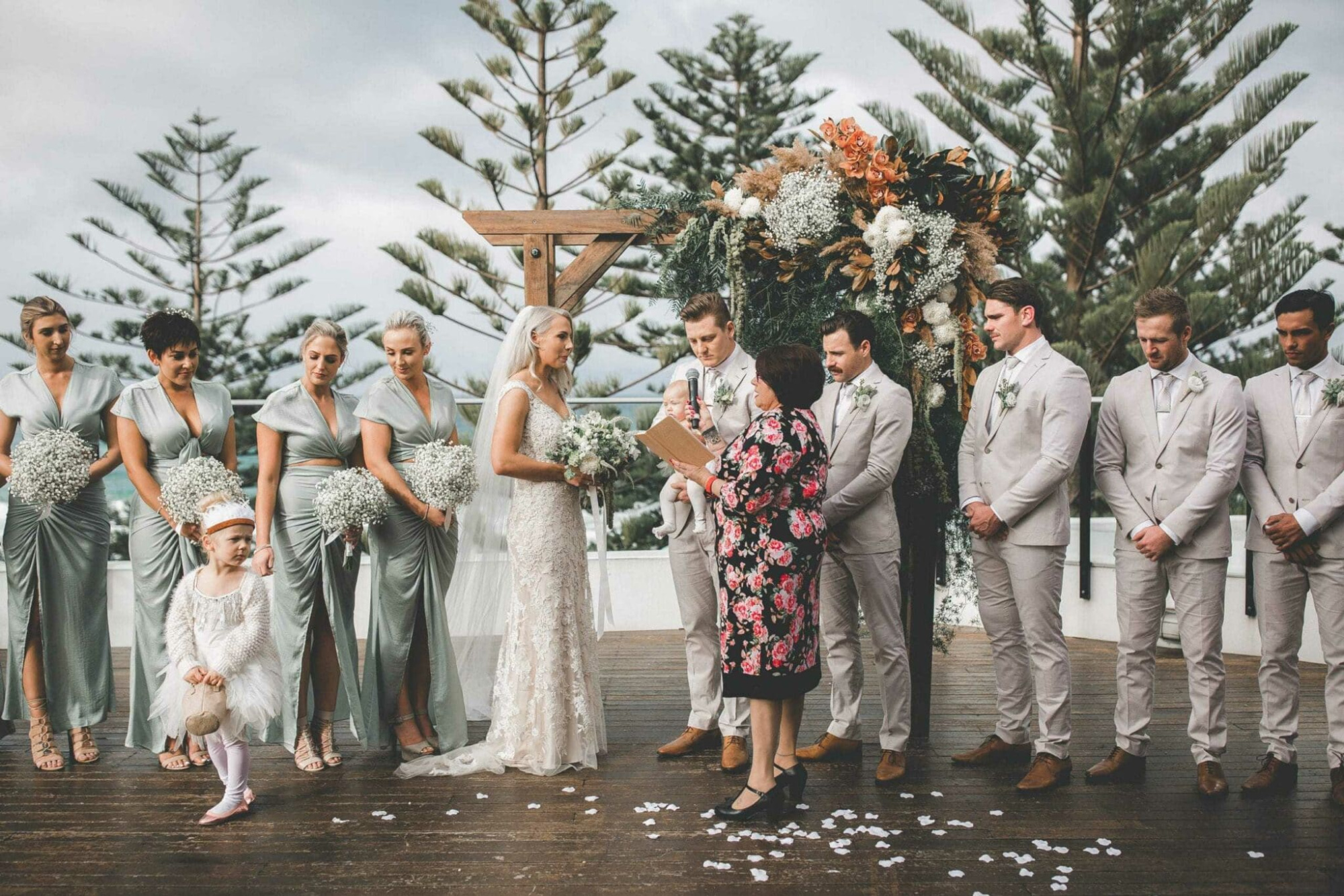 Outdoor wedding ceremony with bride, groom, and bridal party standing before a lush floral arbor.