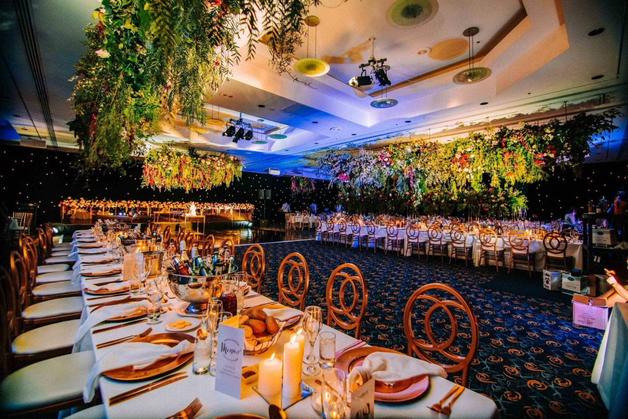 Luxurious indoor wedding reception with long candlelit tables and lush hanging greenery overhead.